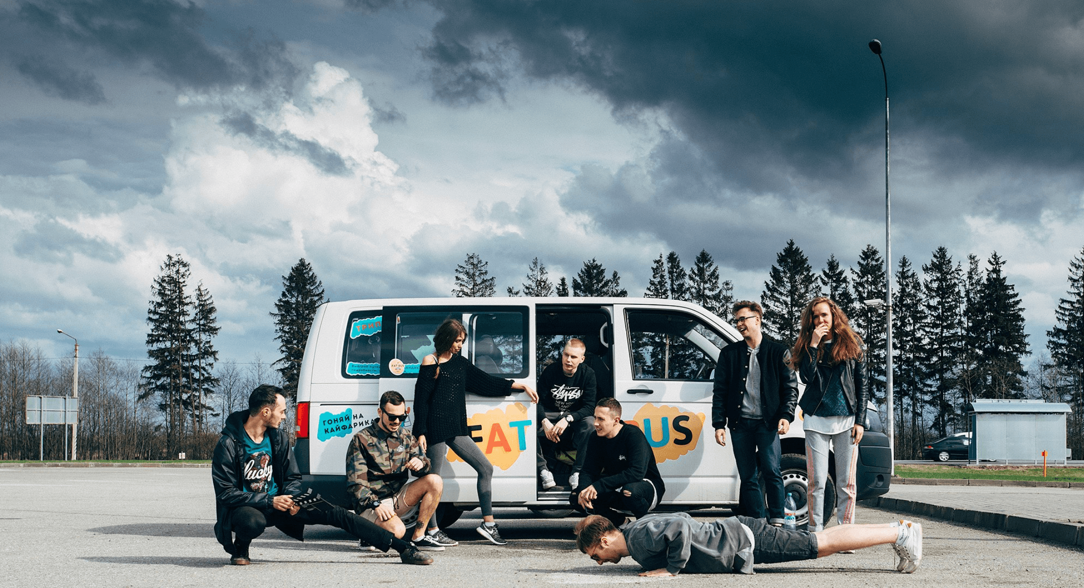 Young people posing around a white van with colorful decals under a dramatic cloudy sky.