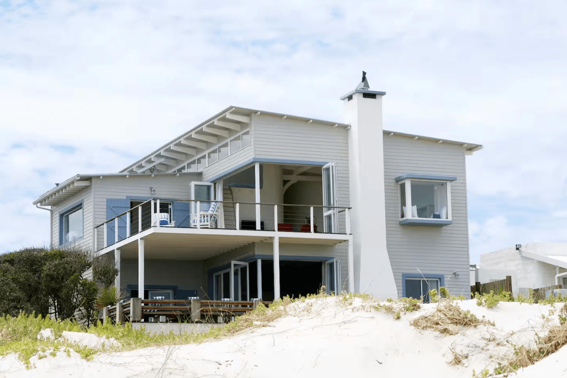Modern grey two-story beach house with white trim and balcony on a sandy dune.
