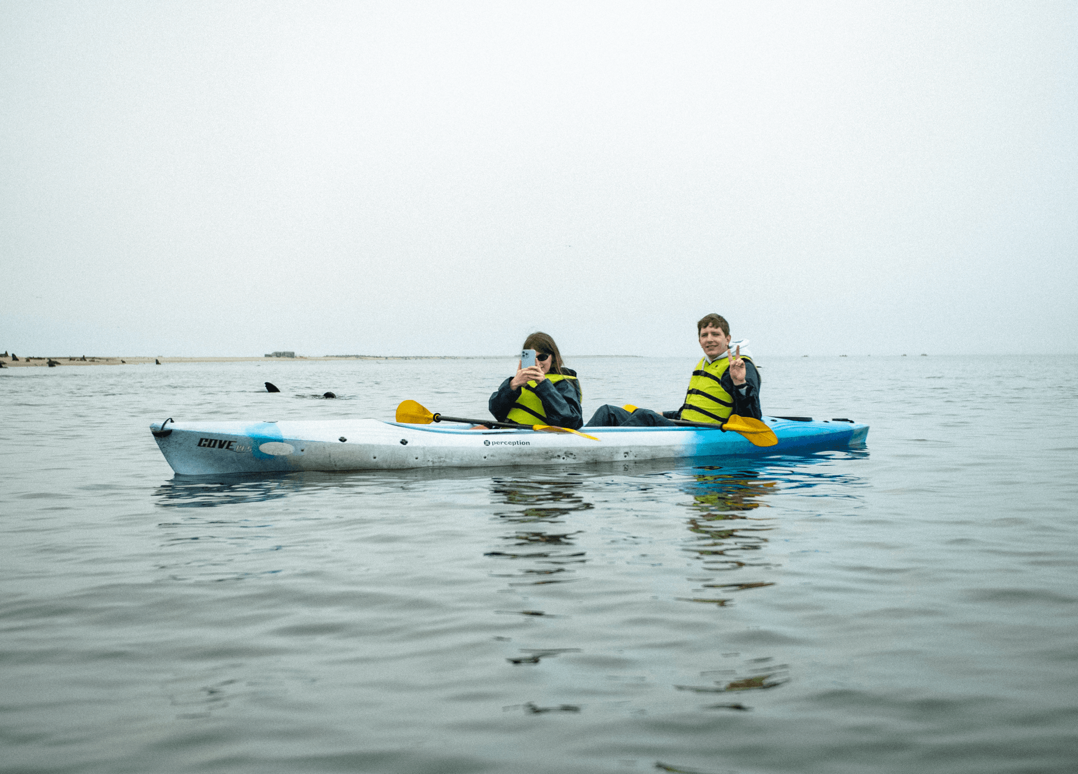 Two people in a tandem kayak on calm water, wearing yellow life vests and posing.