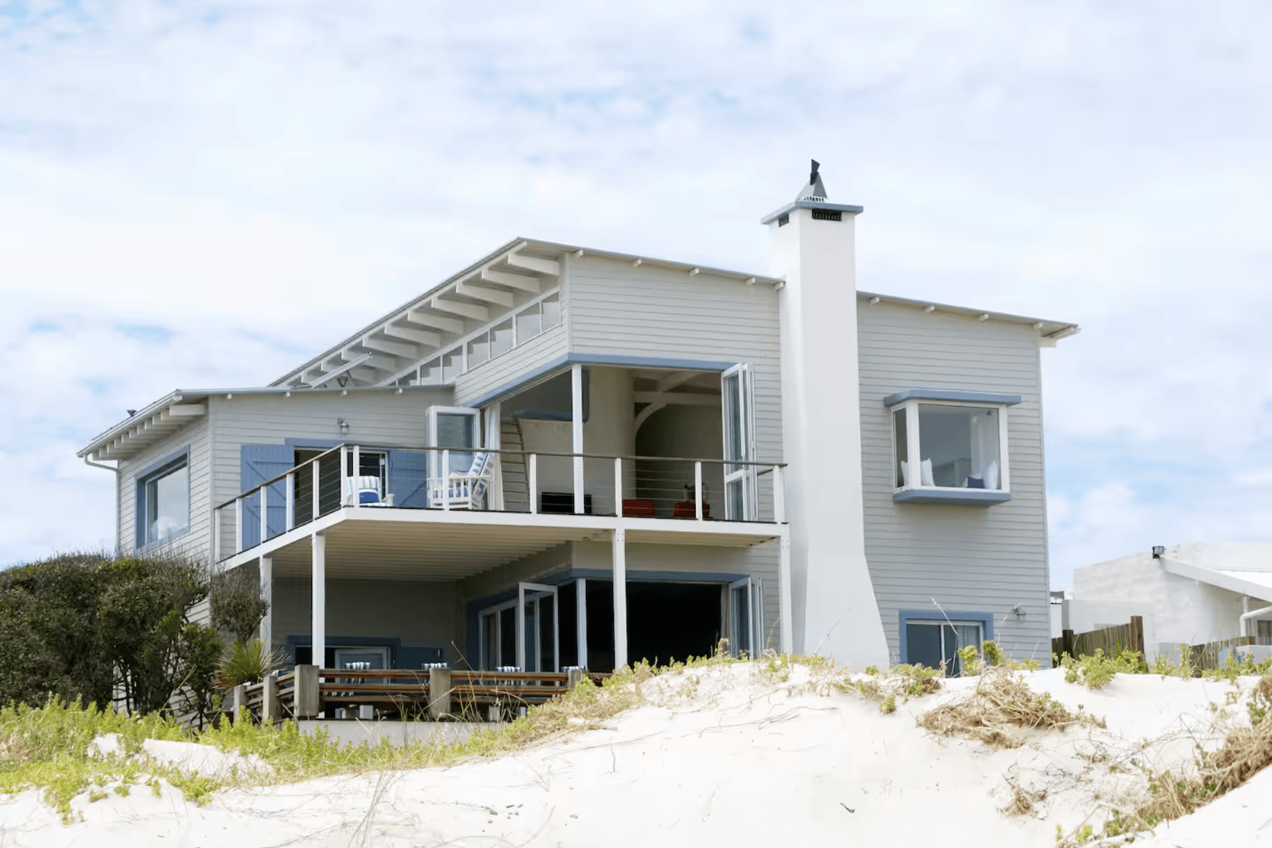 Modern grey beach house with white balconies and a chimney behind a sand dune.