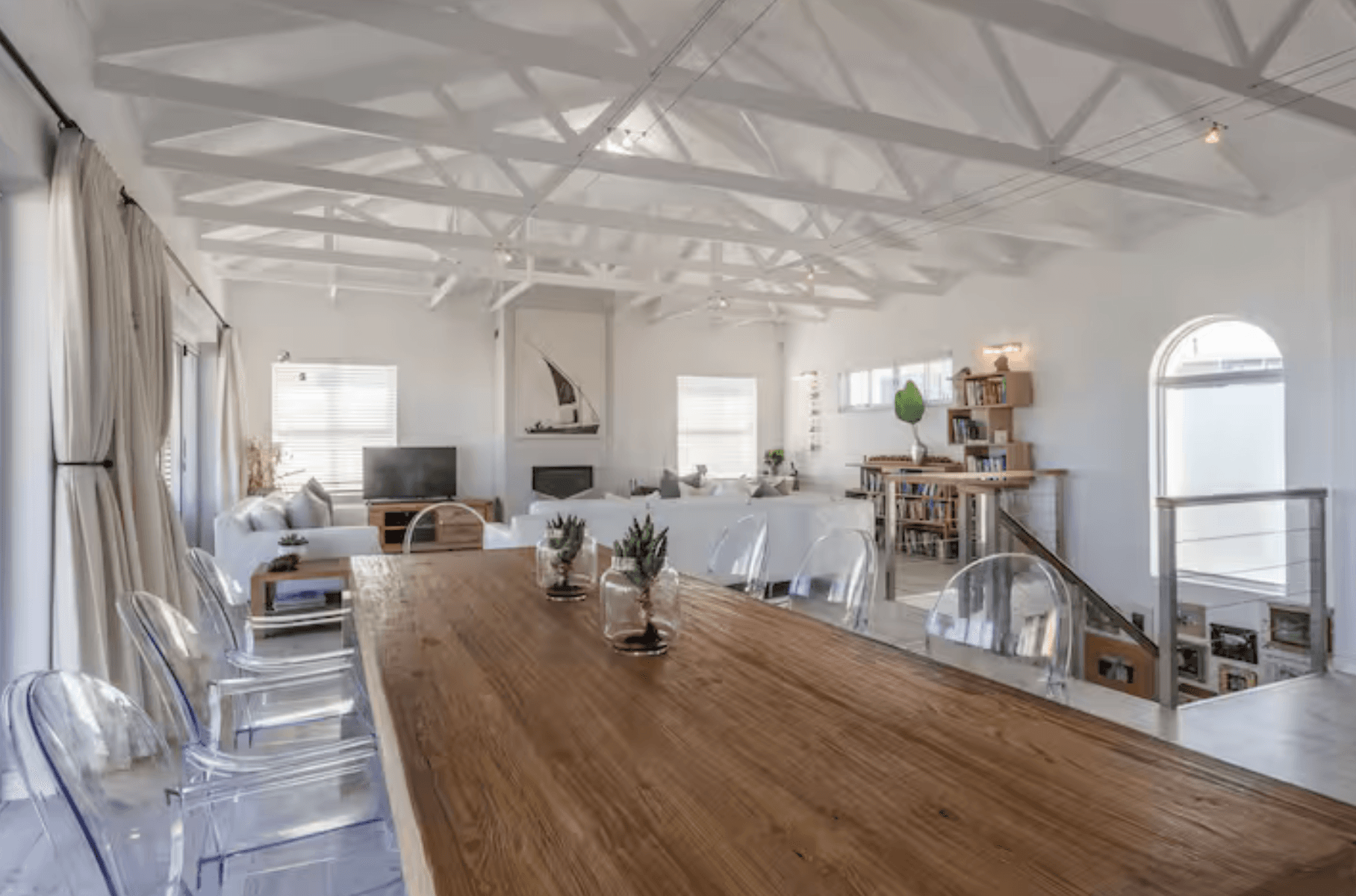 Bright open-plan living space with a long wooden dining table, clear chairs, and vaulted ceilings.