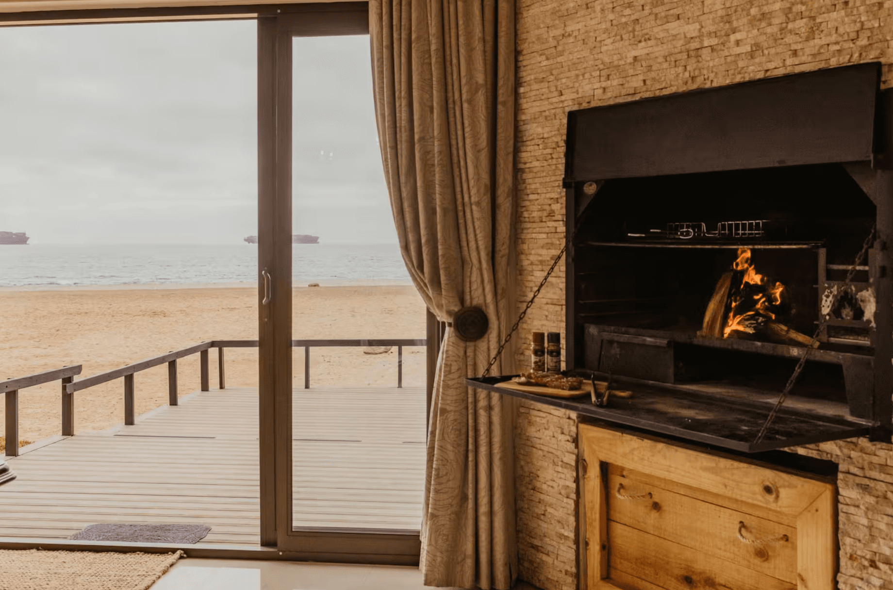 Stone fireplace with a burning fire beside a glass door overlooking the ocean beach.