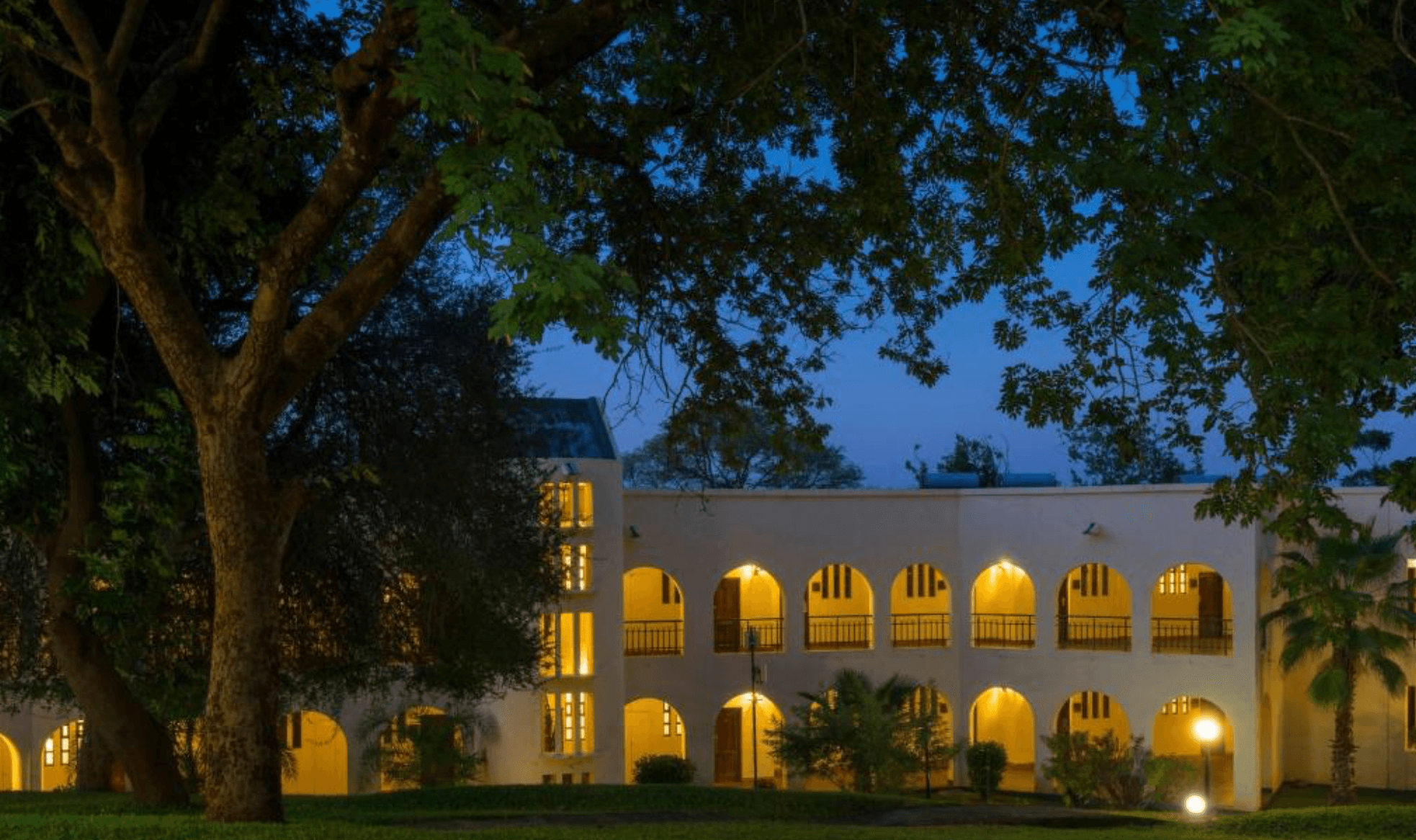 Illuminated white building with arched balconies framed by large leafy trees at dusk.