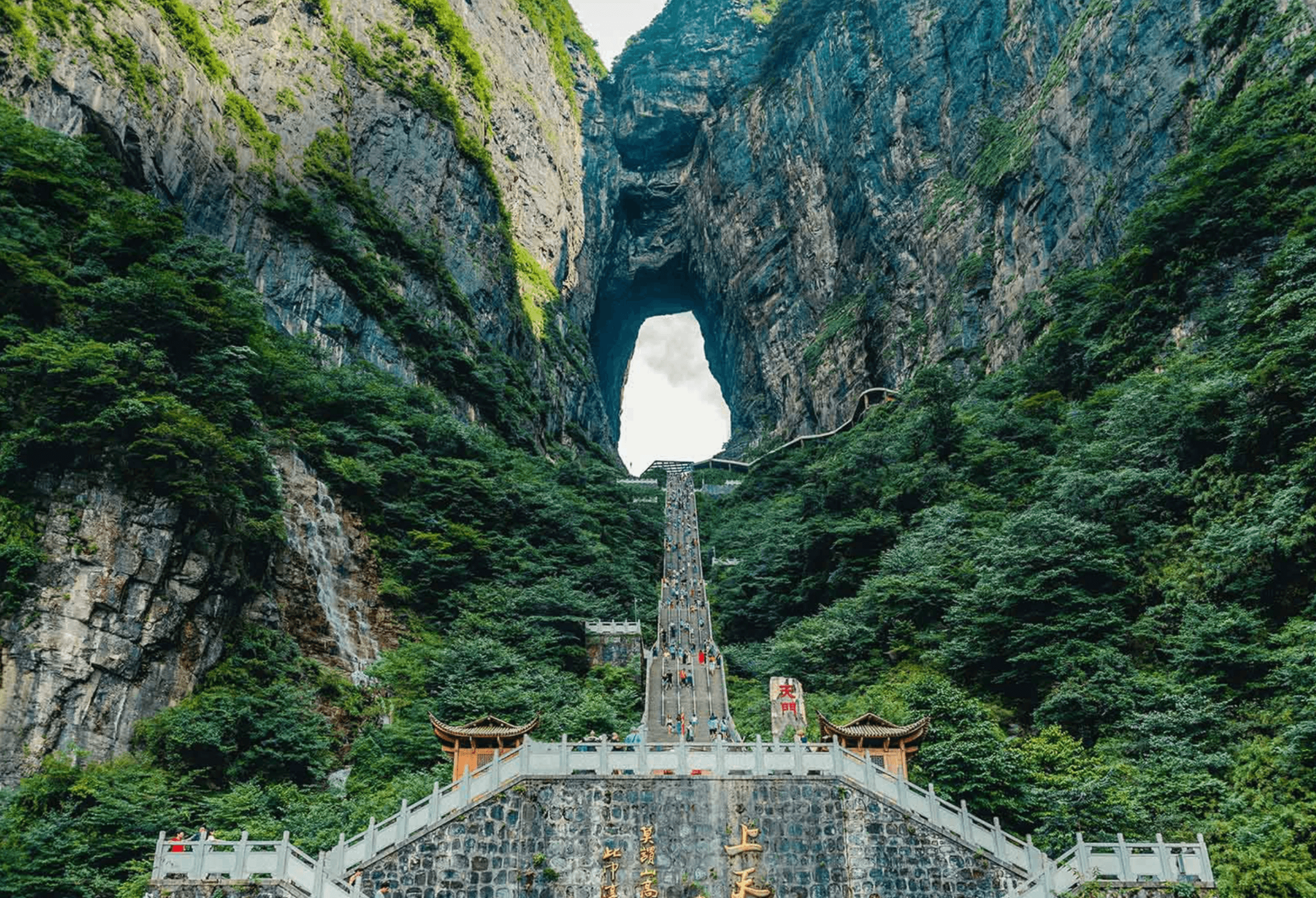 Steep stone stairs leading up to a massive natural archway between lush, towering mountain cliffs.