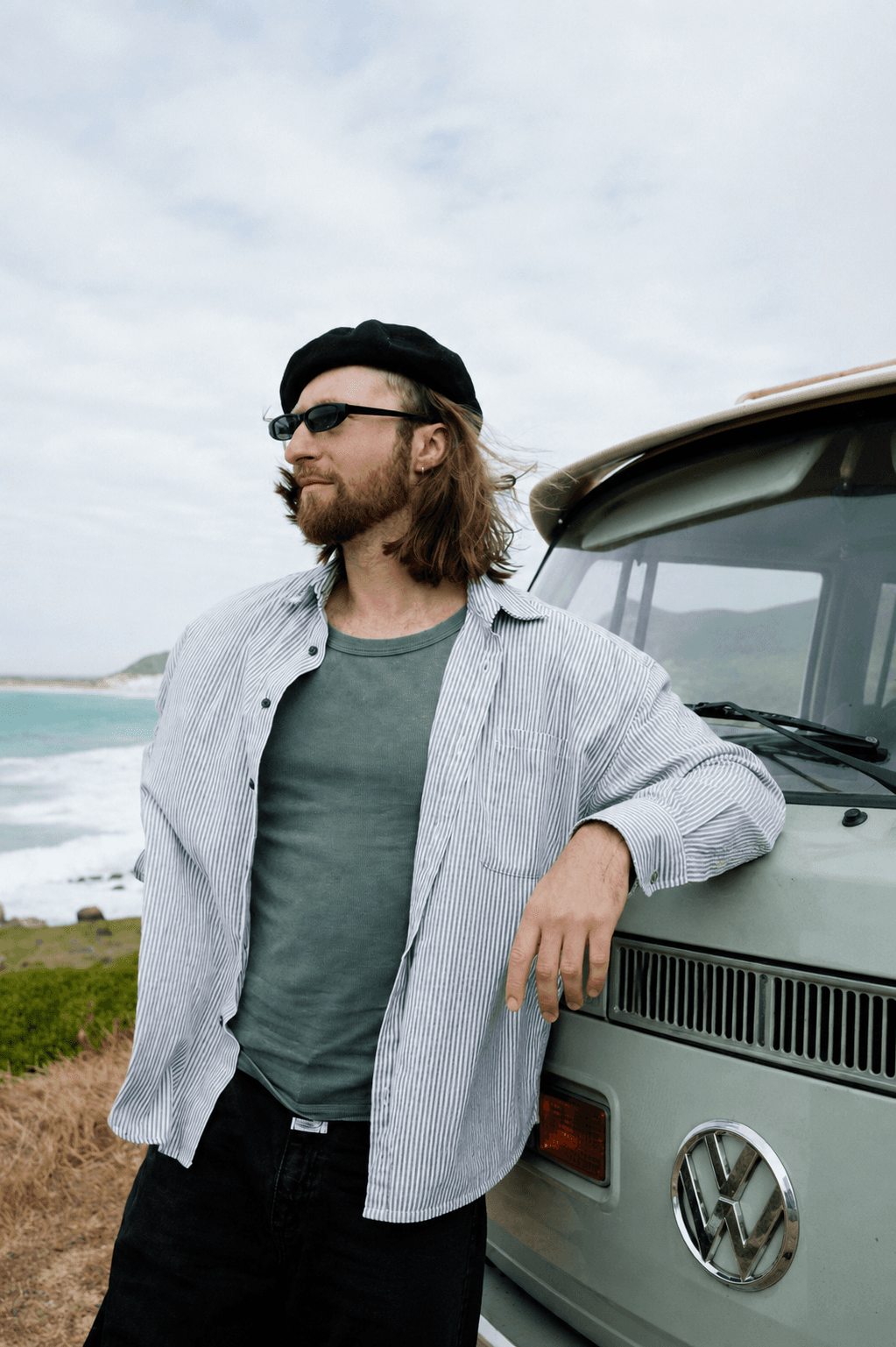 Bearded man in a beret leans against a vintage green van overlooking a coastal landscape.