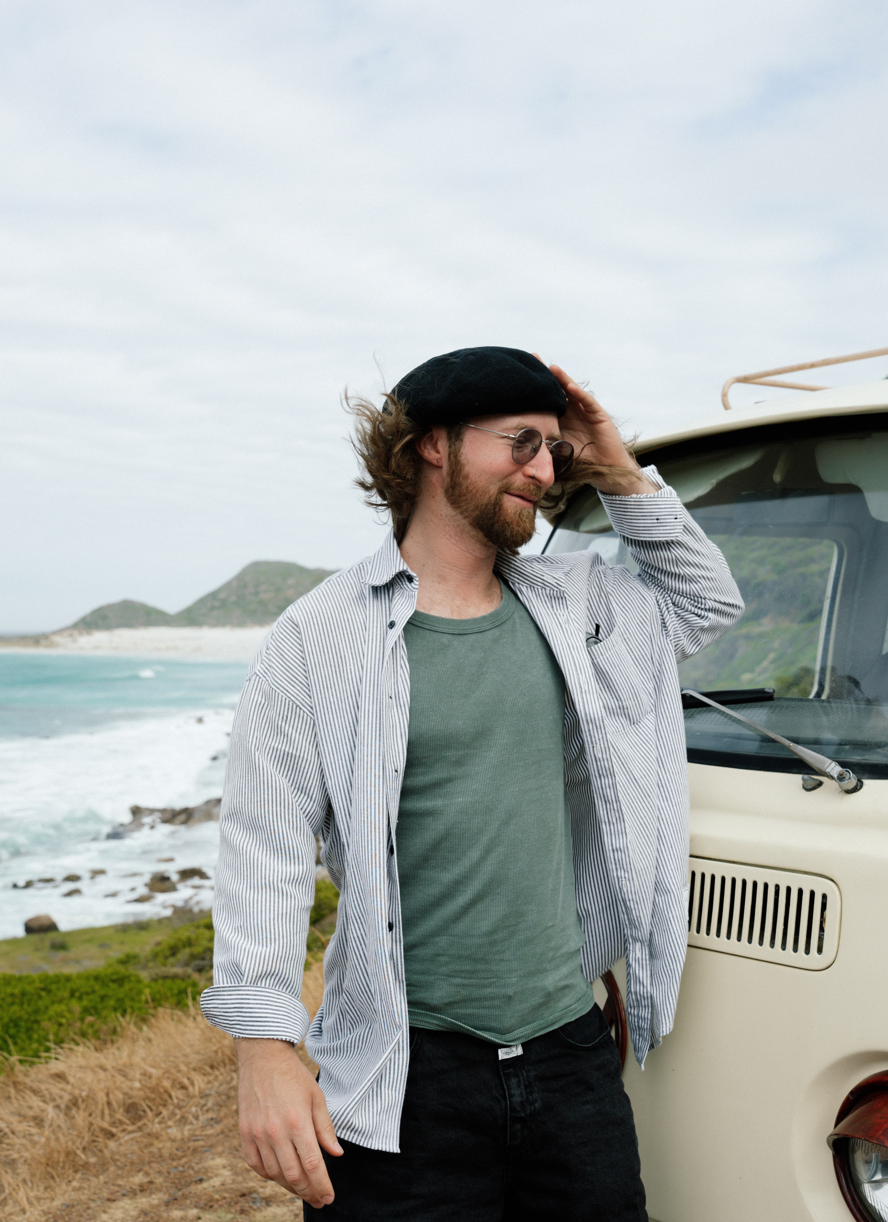 Bearded man in a black beret stands beside a vintage van overlooking a scenic coast.
