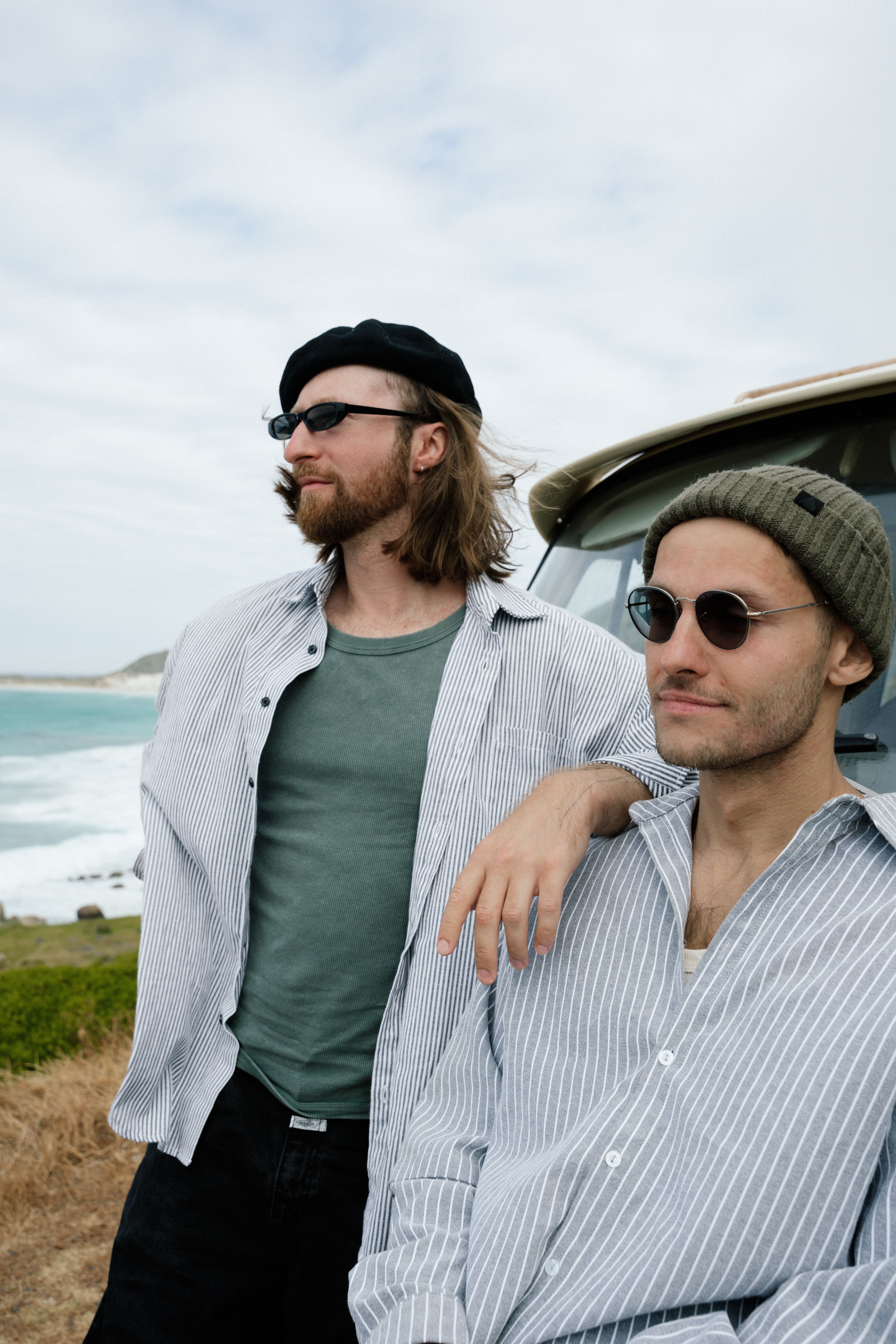Two men wearing sunglasses and striped shirts stand near a van overlooking the ocean.
