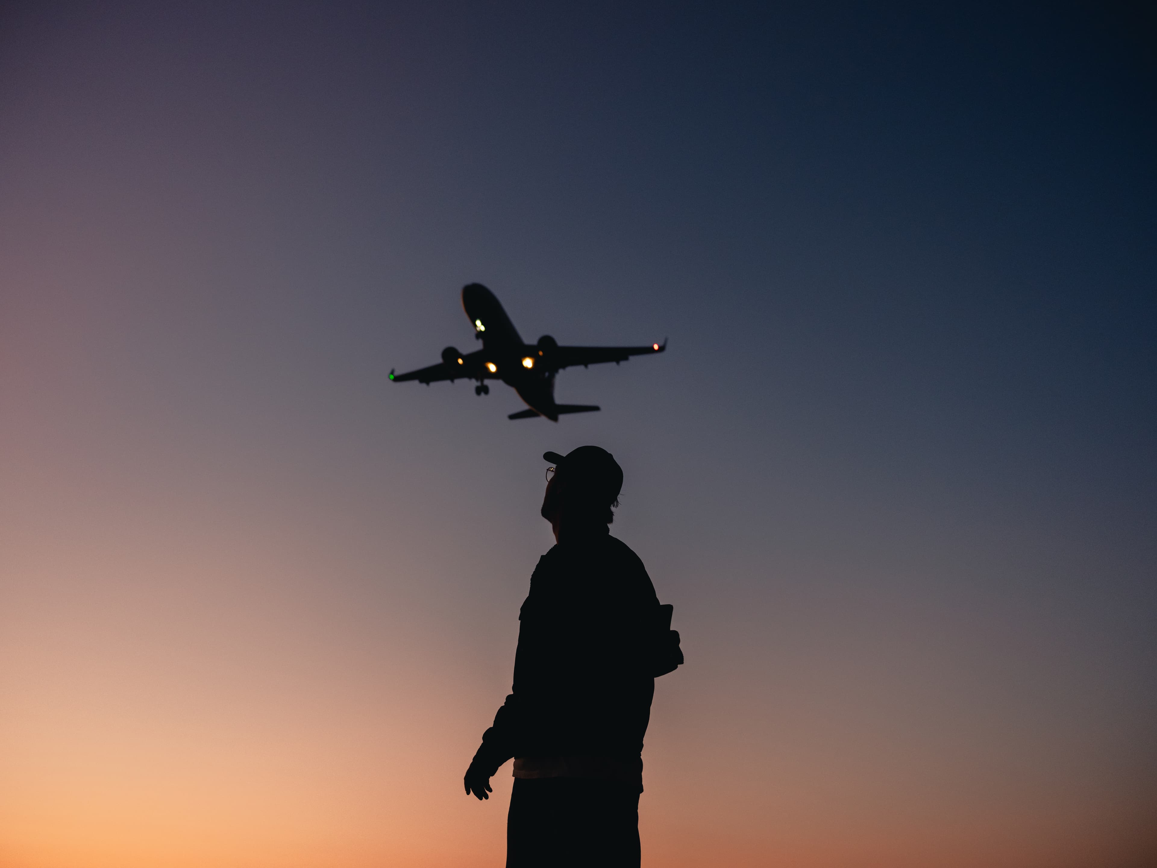 Silhouette of a person looking up at an airplane flying overhead during a colorful sunset.