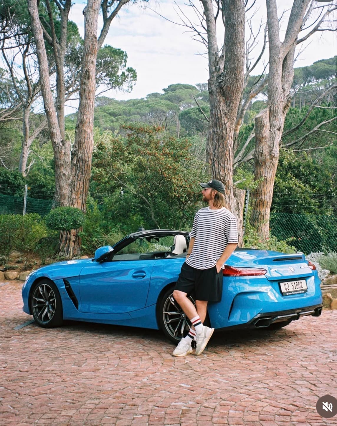 Man in striped shirt leans against blue BMW convertible on cobblestone driveway near tall trees.