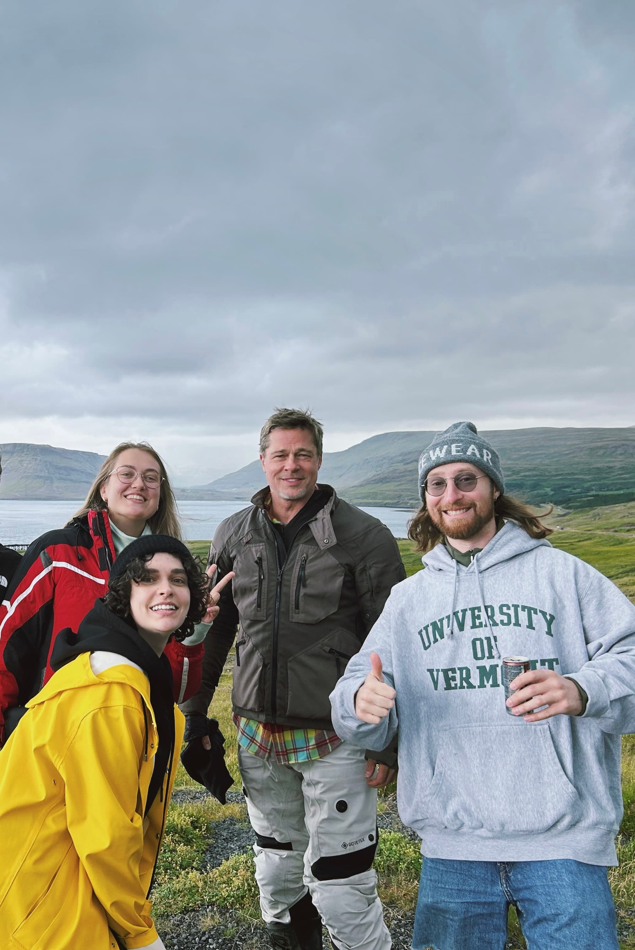 Brad Pitt and three fans pose in a scenic coastal landscape under a cloudy sky.