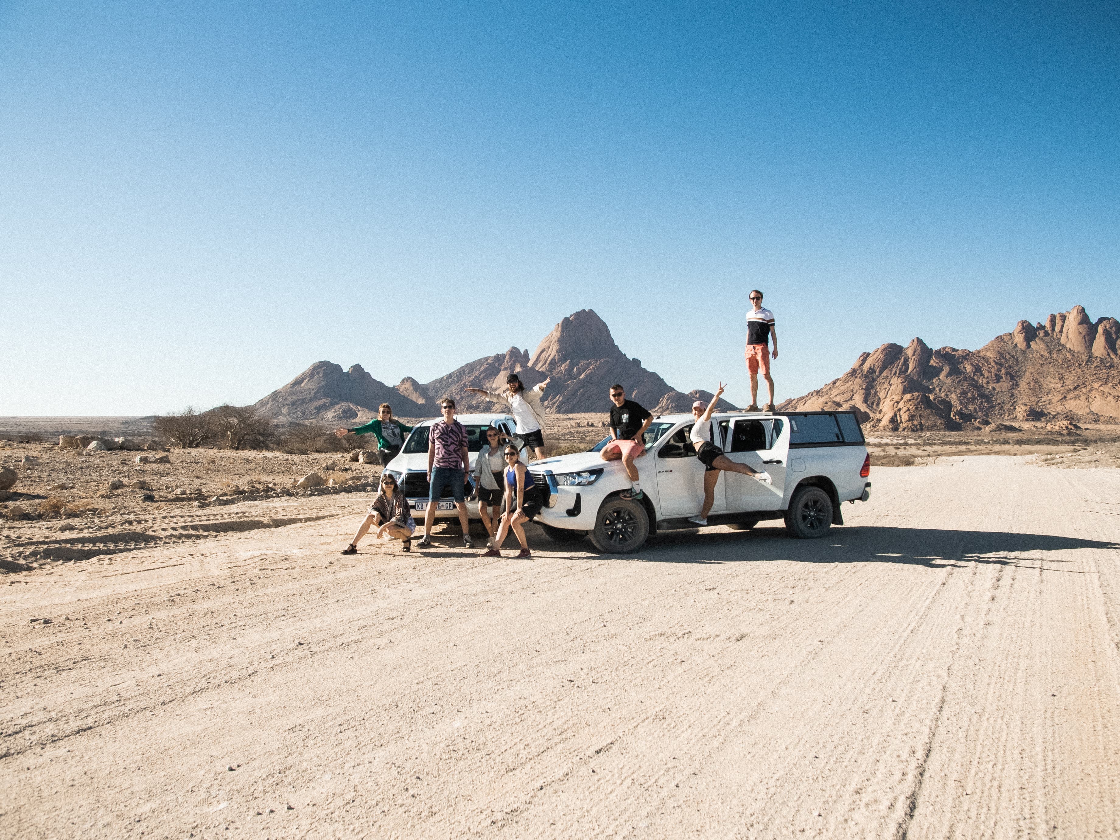 Friends posing with white pickup trucks in a desert landscape with large rocky mountains.