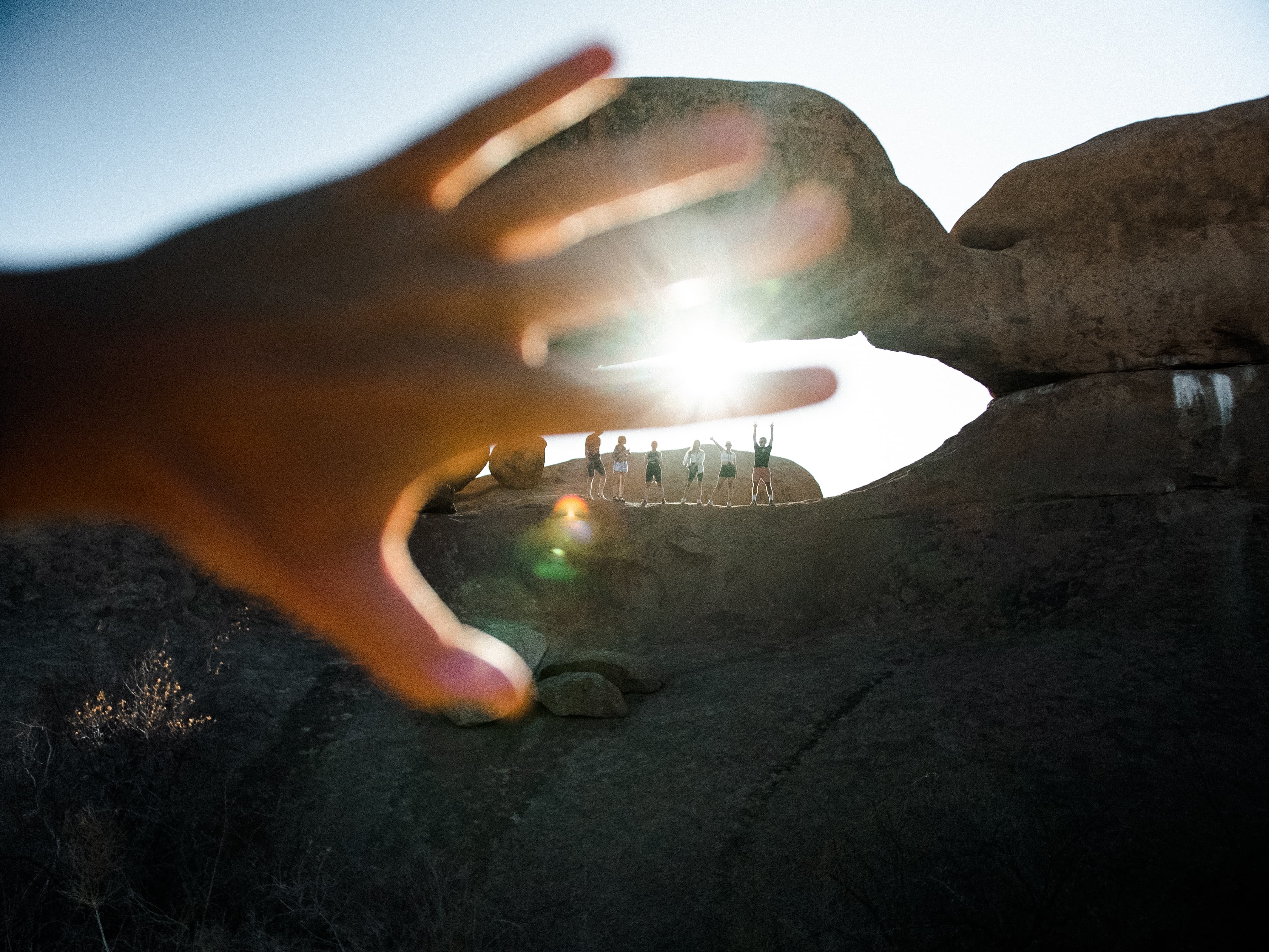 A blurry hand reaches toward silhouetted people standing in a rock arch under bright sun.