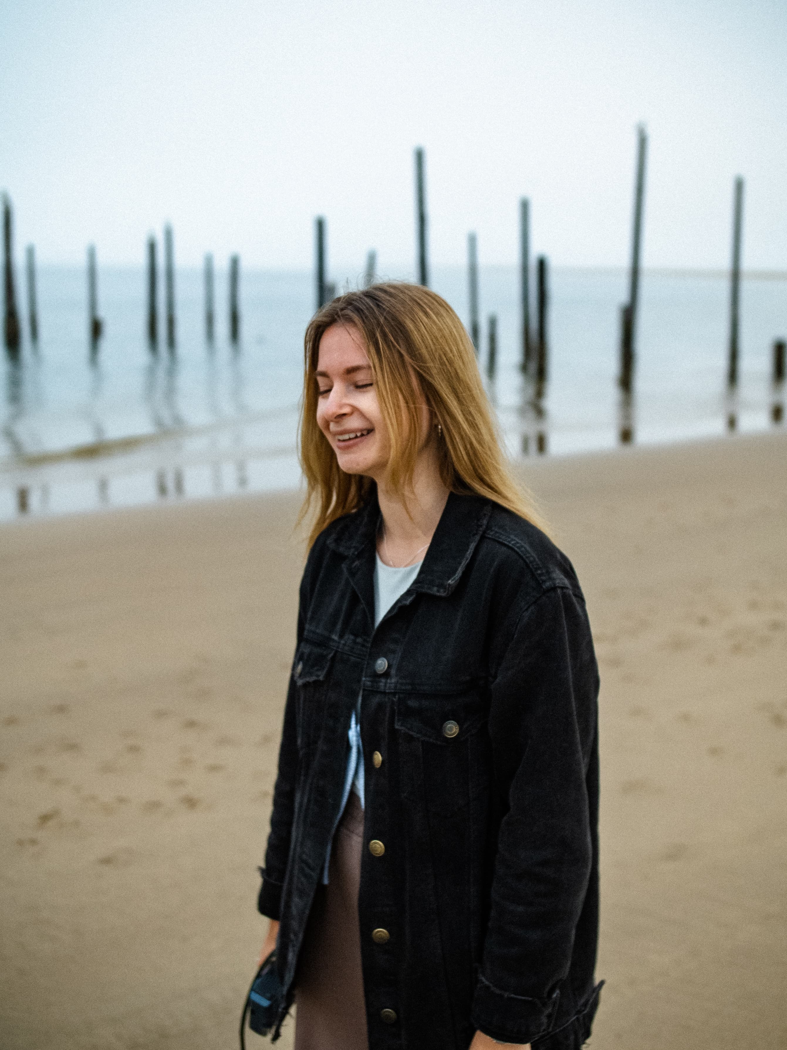 Smiling blonde woman in a black denim jacket on a sandy beach with wooden pilings.