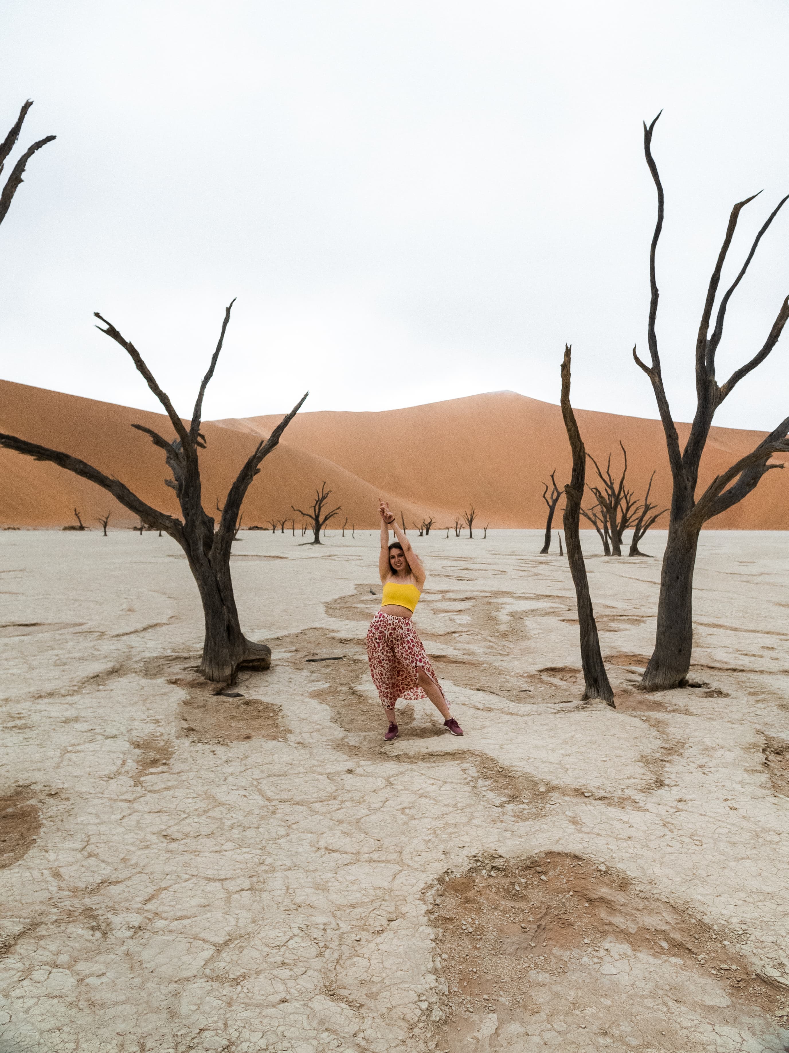 Woman posing among dead trees on a white clay pan with orange sand dunes.