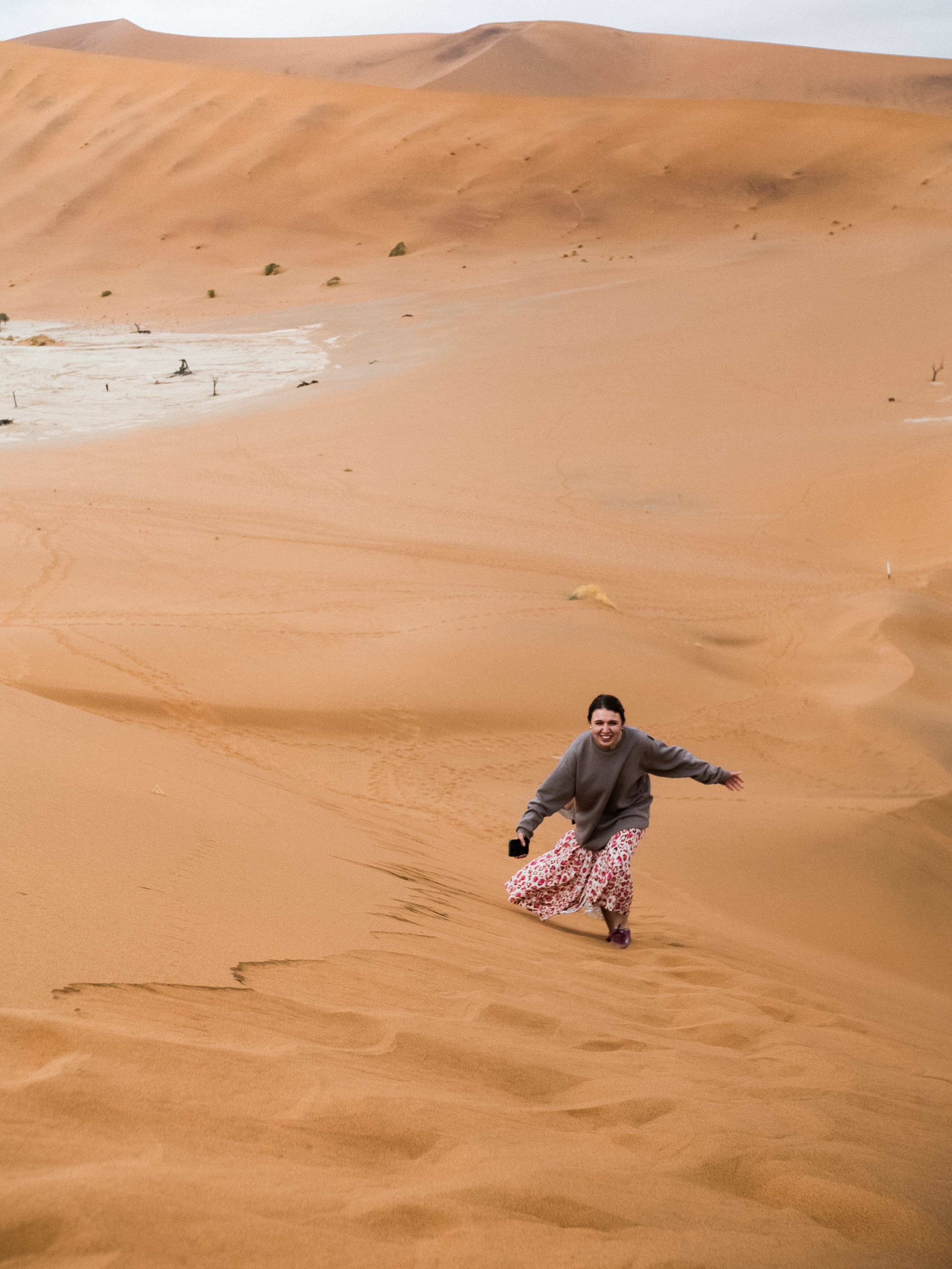 Smiling woman in a long skirt runs down a massive orange sand dune.