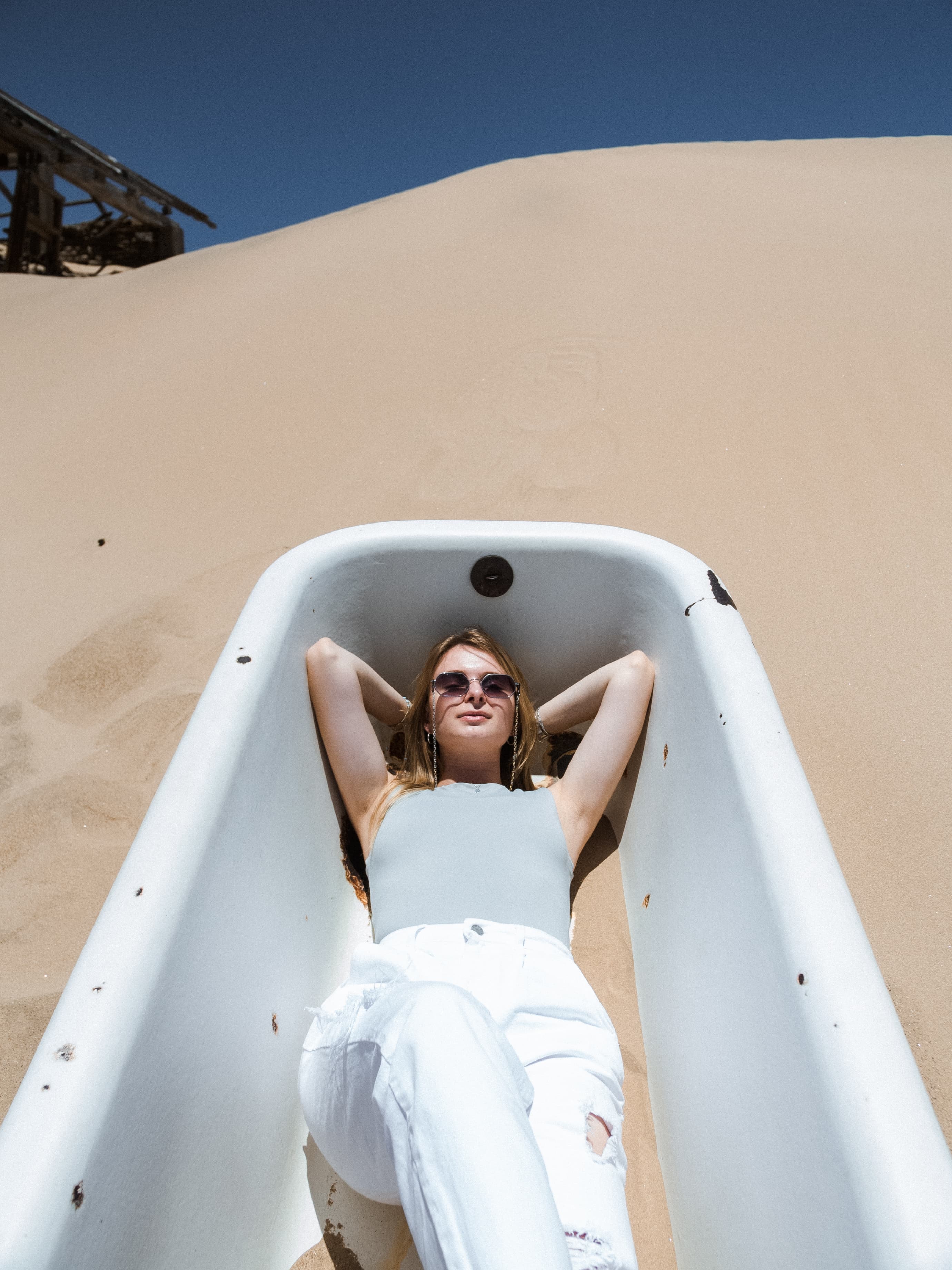 Woman in sunglasses relaxes in a white bathtub nestled within a vast desert sand dune.