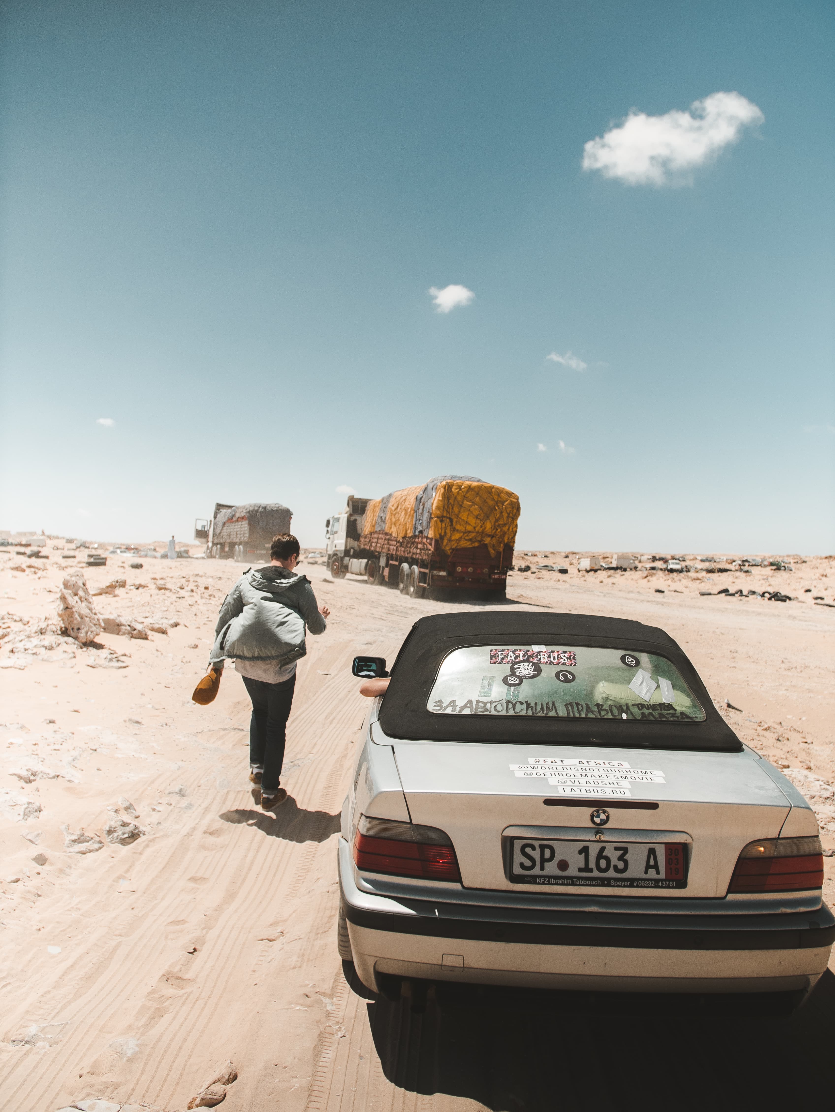 Silver convertible on a desert road with a person walking toward distant, loaded trucks.