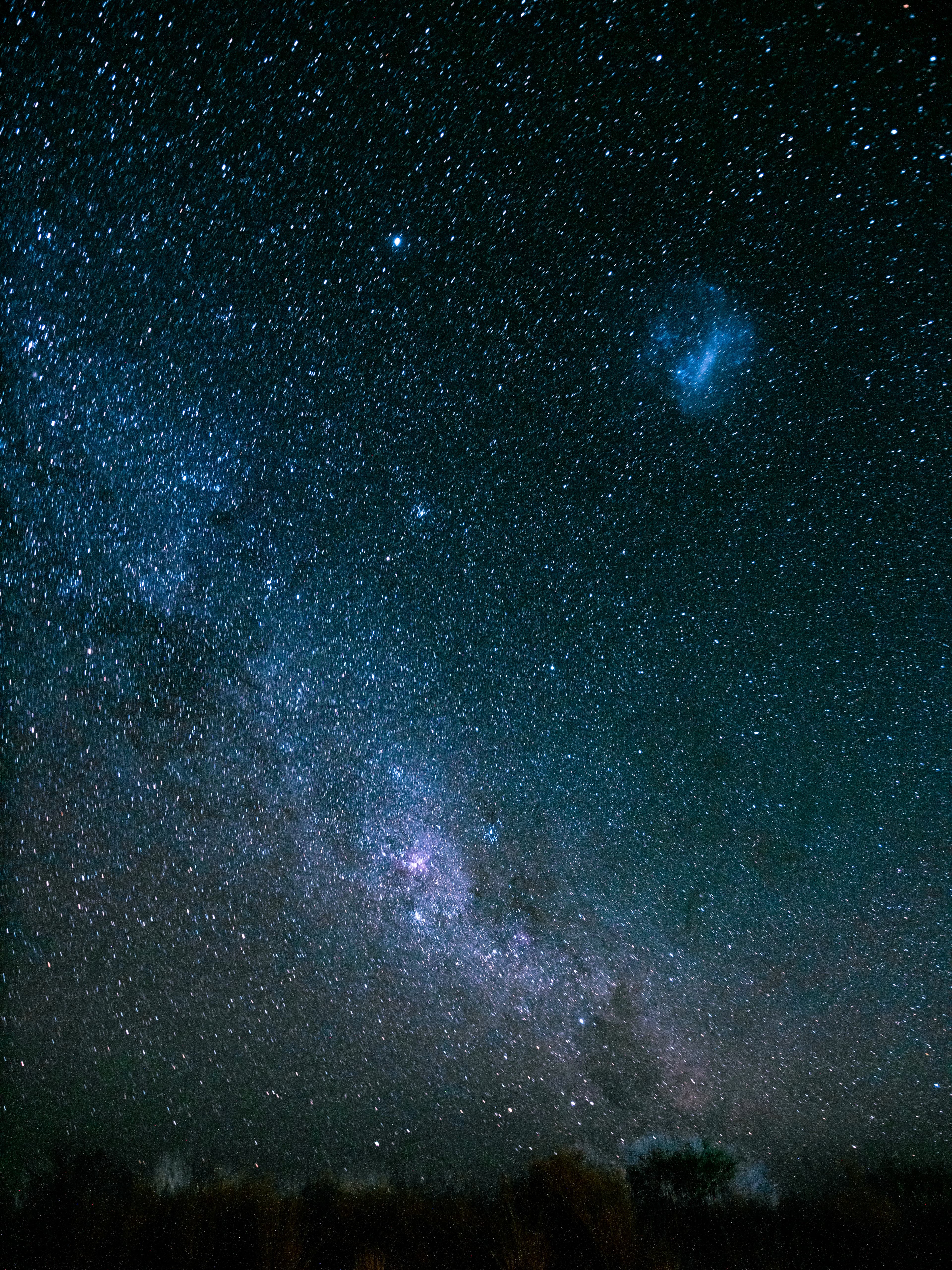 A vibrant night sky with the Milky Way and a blue nebula over dark silhouettes.