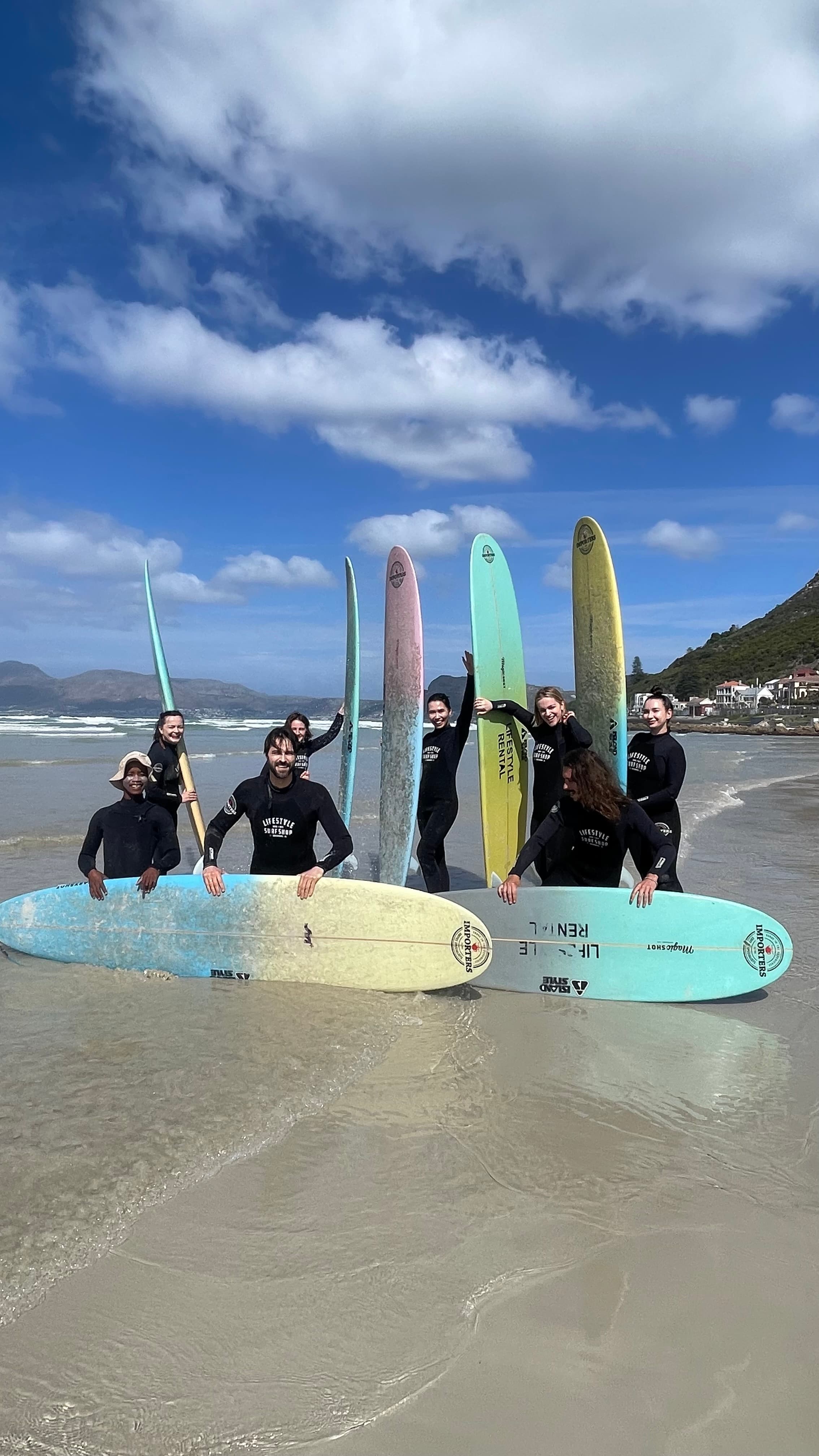 Group of surfers in black wetsuits posing with colorful surfboards on a sunny beach.