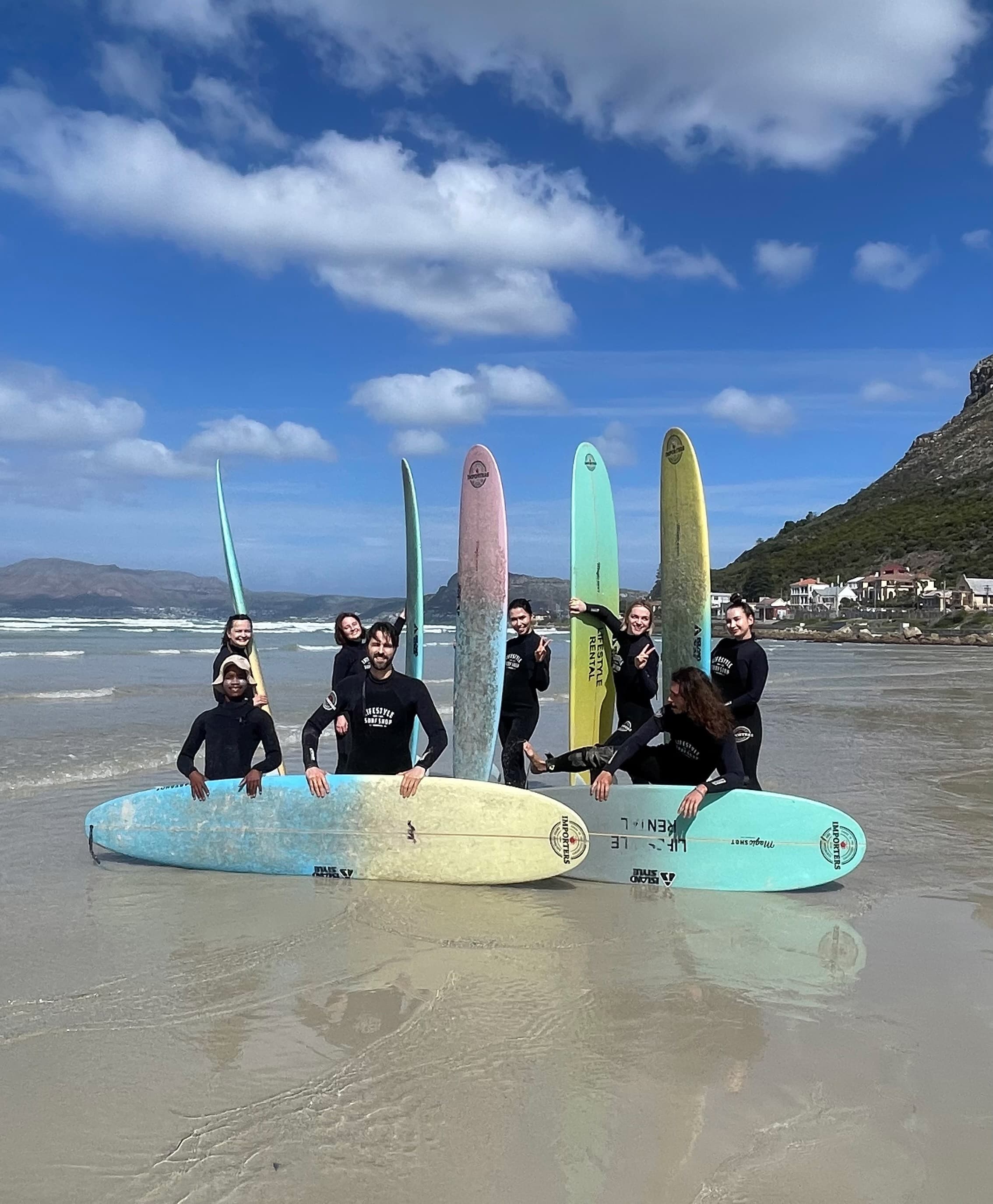 Seven surfers in wetsuits pose with colorful surfboards on a sunny beach with distant mountains.