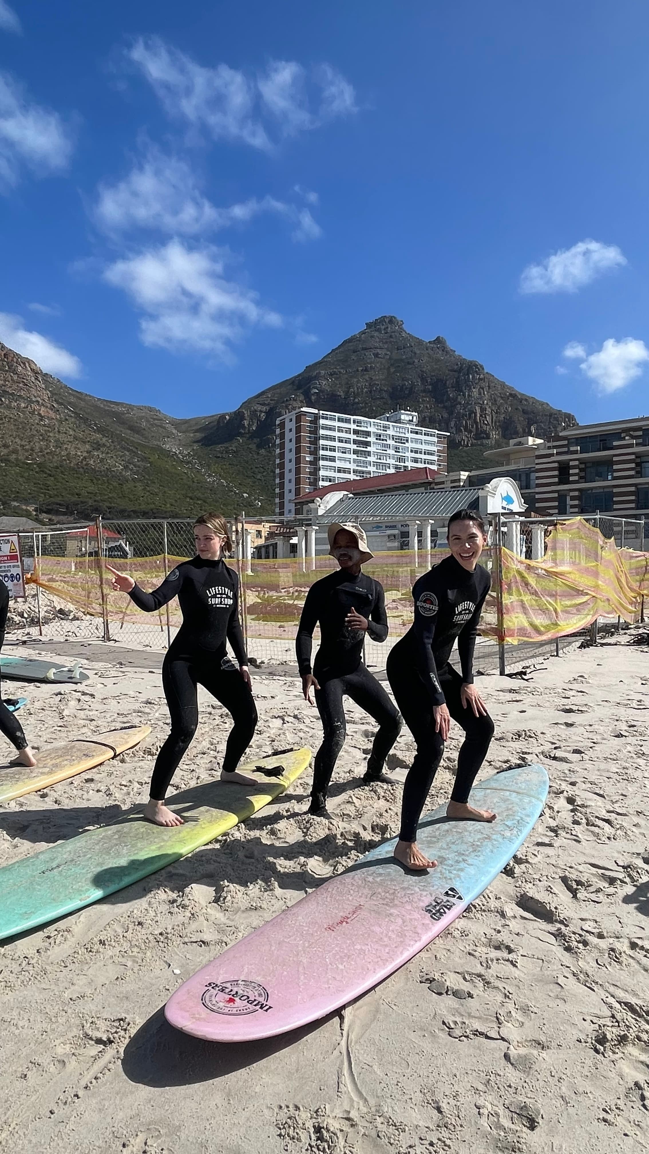 Three people in wetsuits practice surfing on boards on a beach with a mountain backdrop.