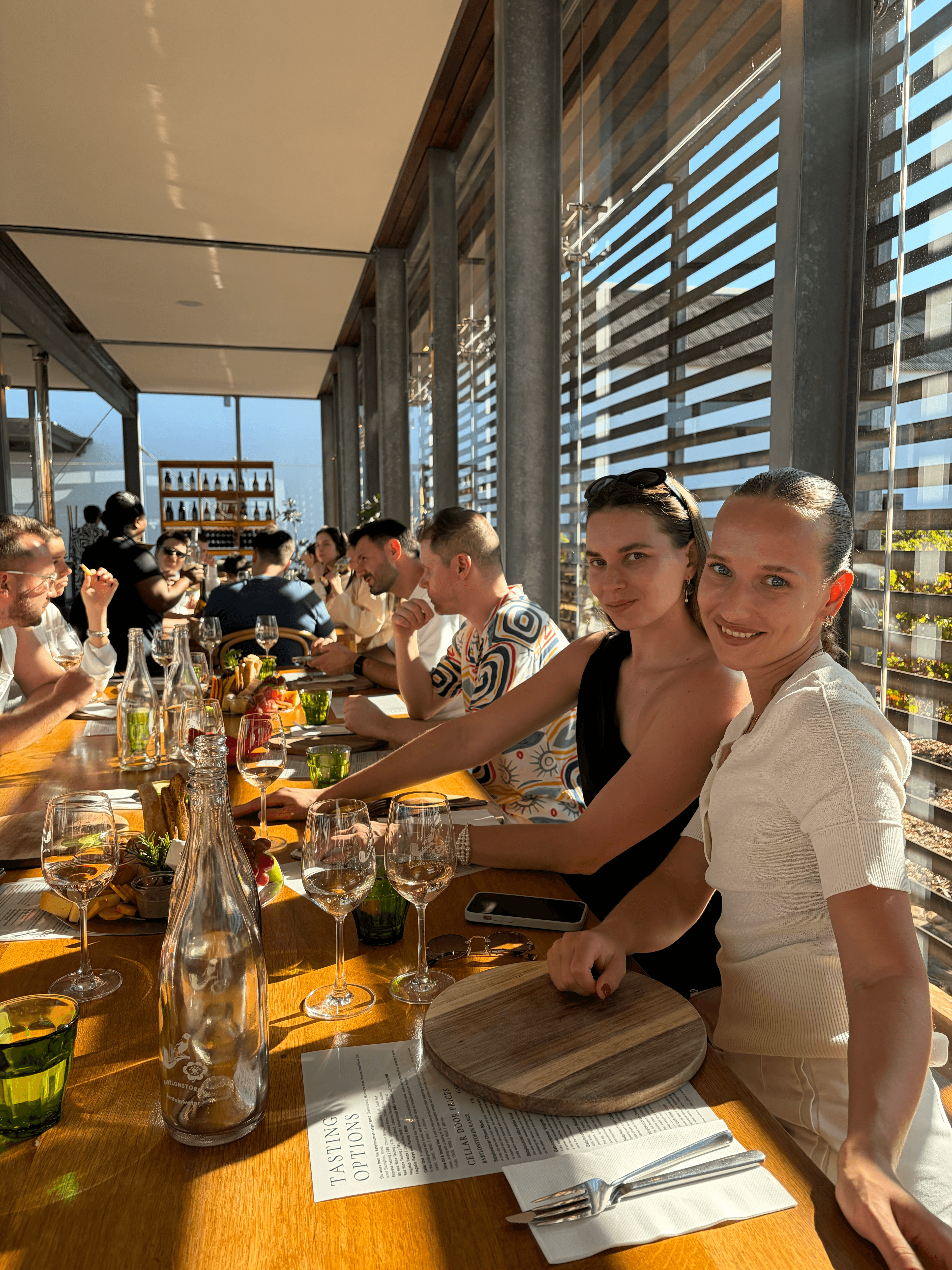 Two women smile at a long table with wine and food in a sunlit restaurant.