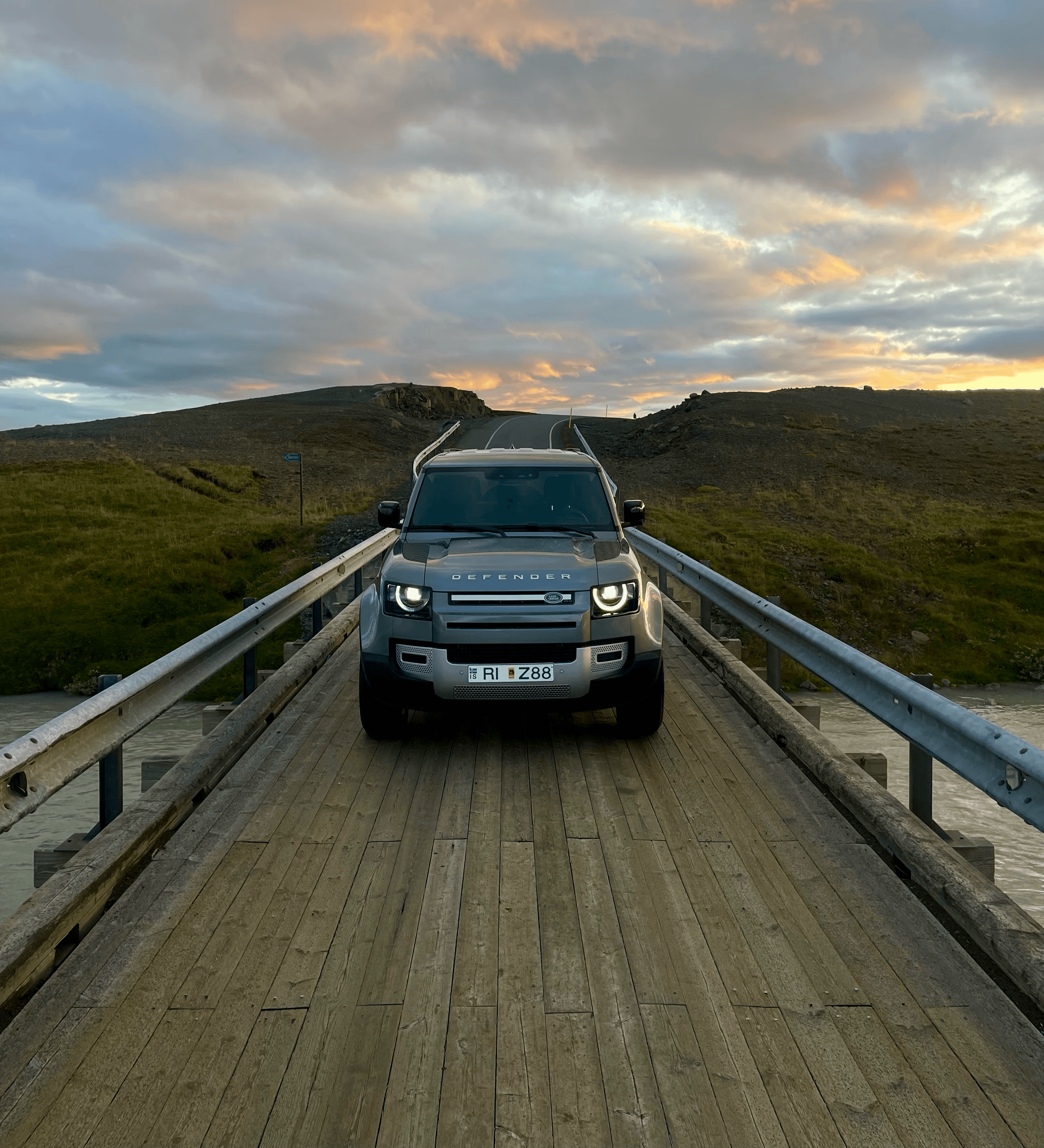 Silver Land Rover Defender parked on a wooden bridge under a colorful sunset sky.