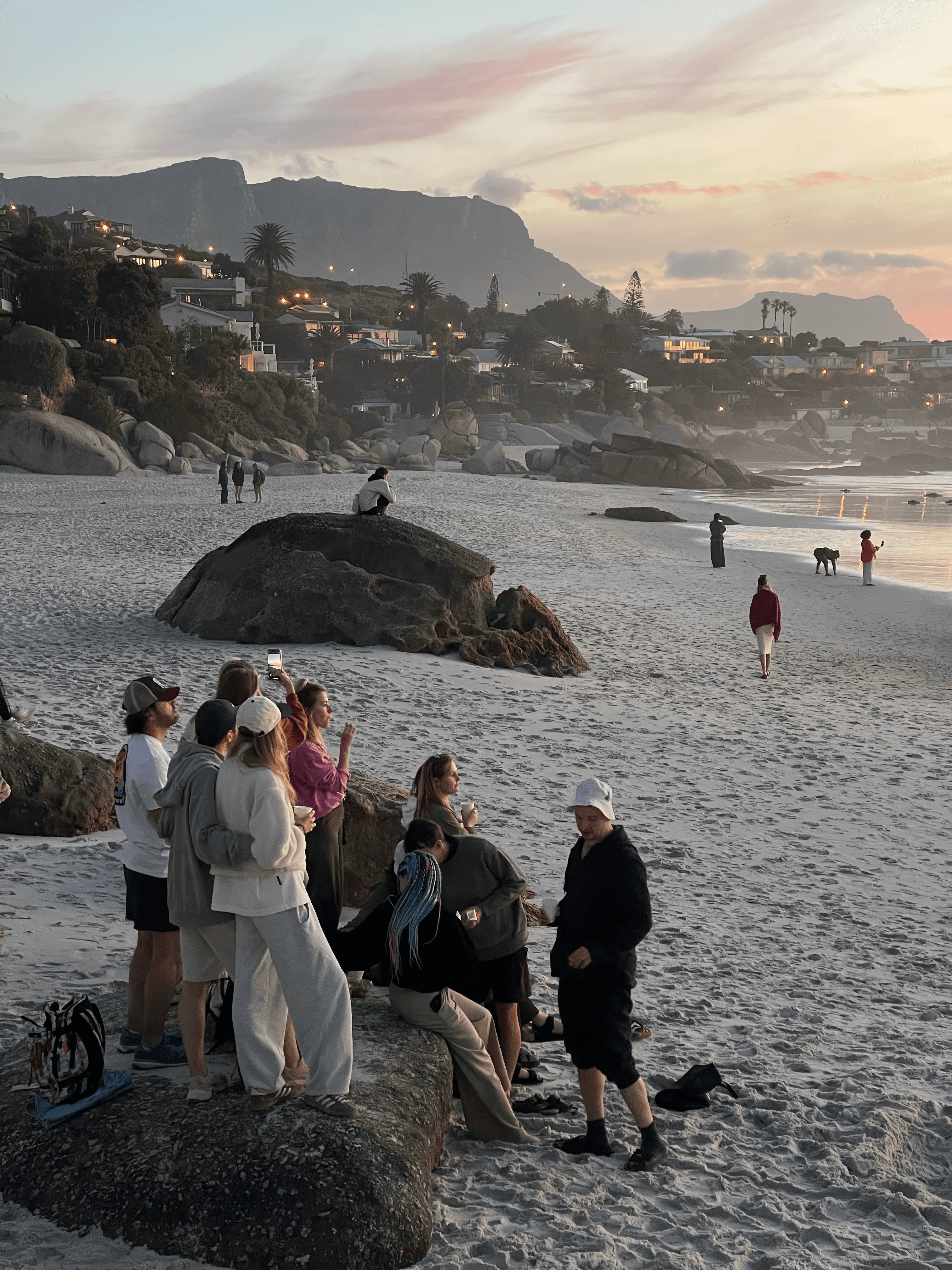 People gathering on a sandy beach at sunset with large boulders and distant mountains.