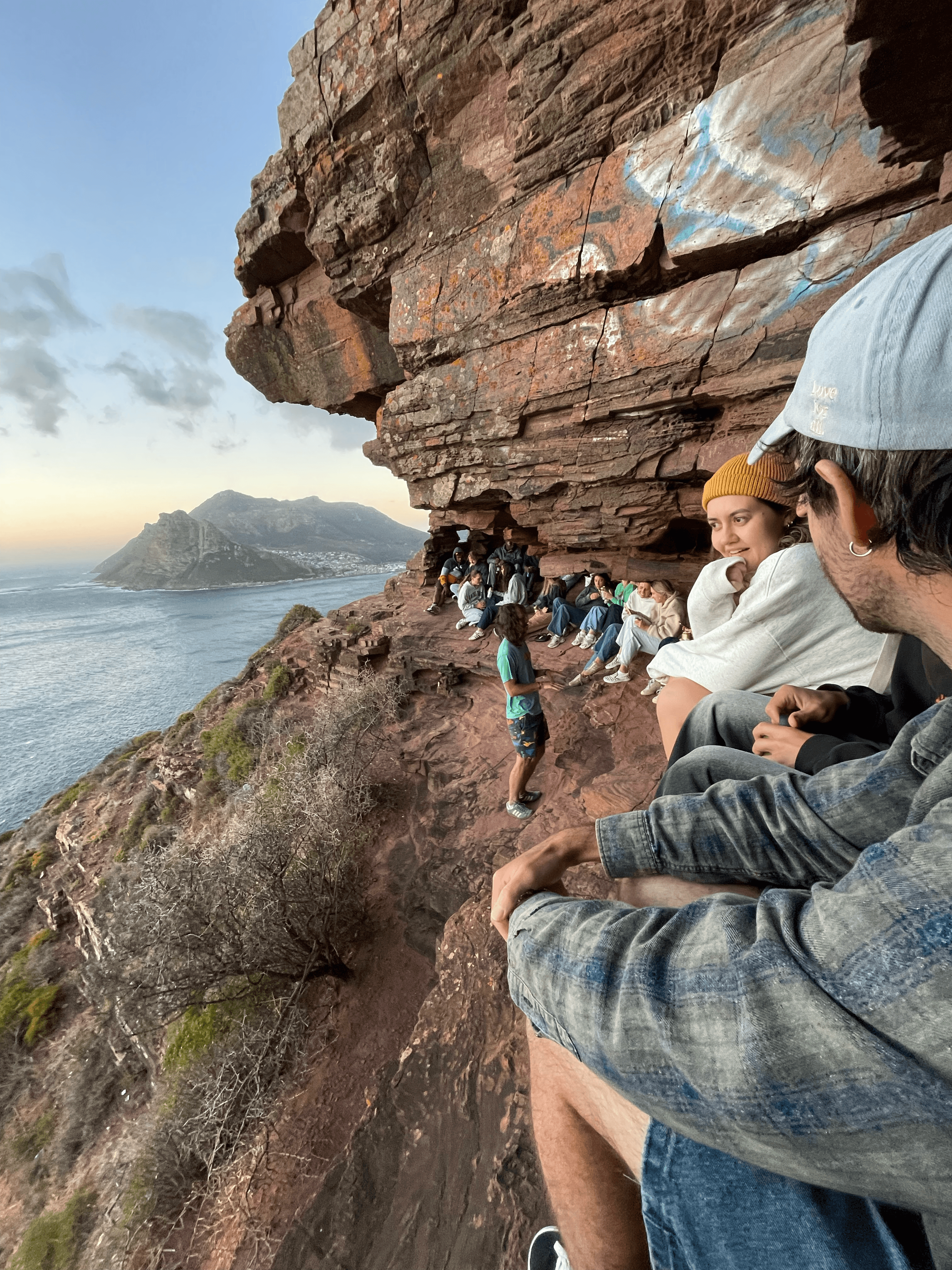 Group of friends sit on a rocky ledge overlooking a scenic coastal mountain and ocean.