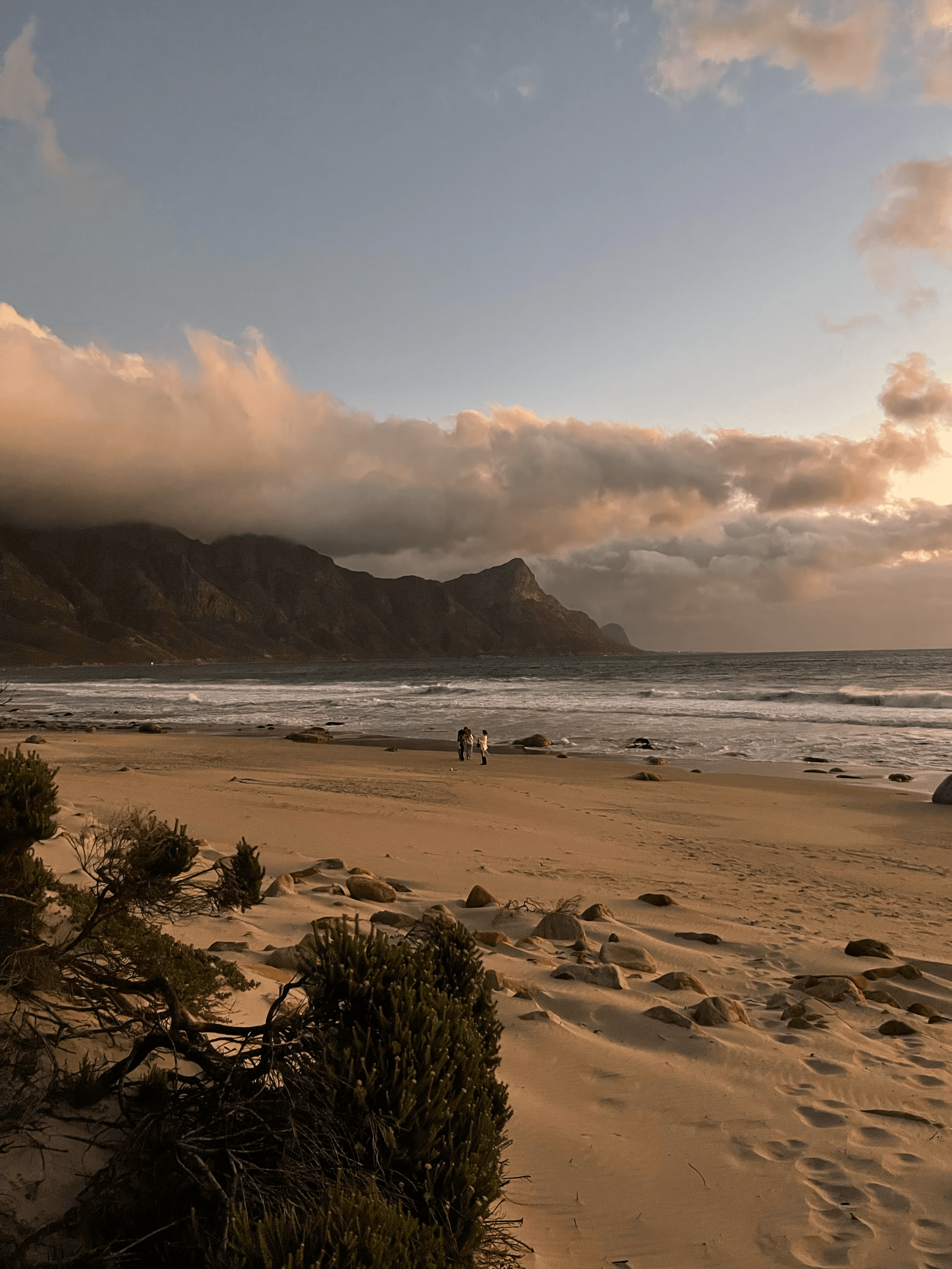 Sandy beach at sunset with mountains under thick clouds and people walking near the waves.