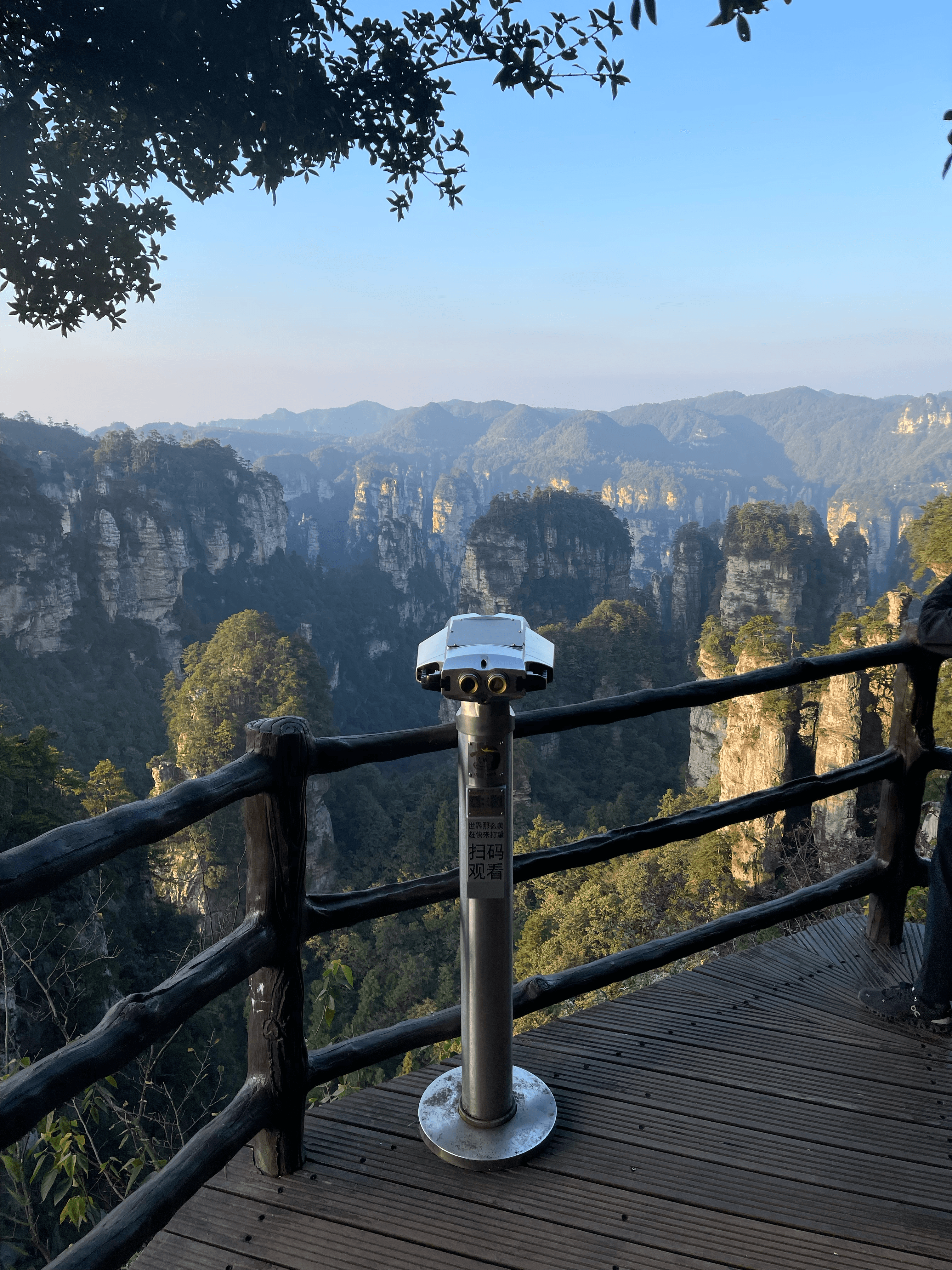 Binoculars on a wooden platform overlook a lush mountain valley of towering rock pillars.