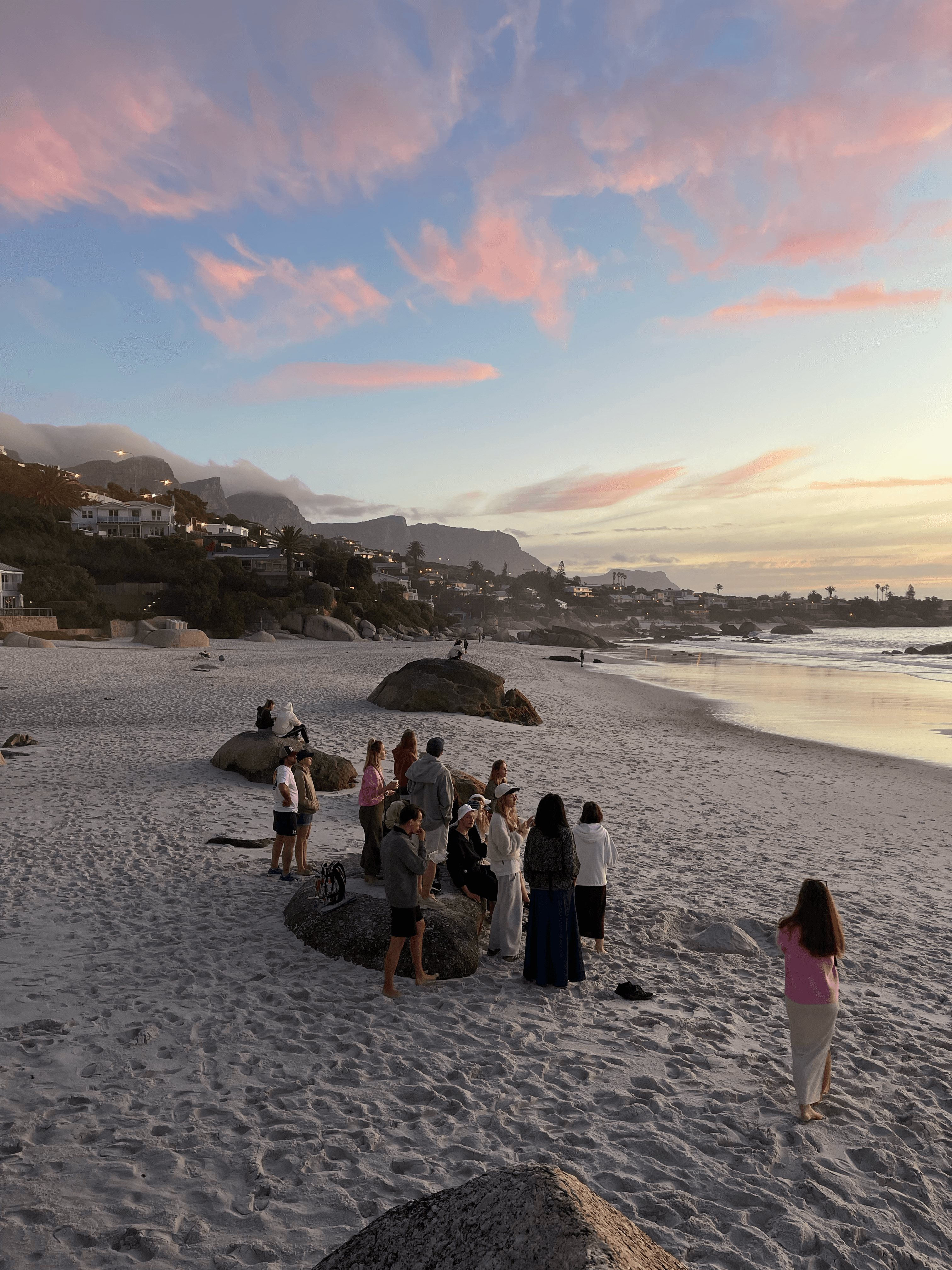 People gather on a white sand beach with boulders under a vibrant pink sunset sky.