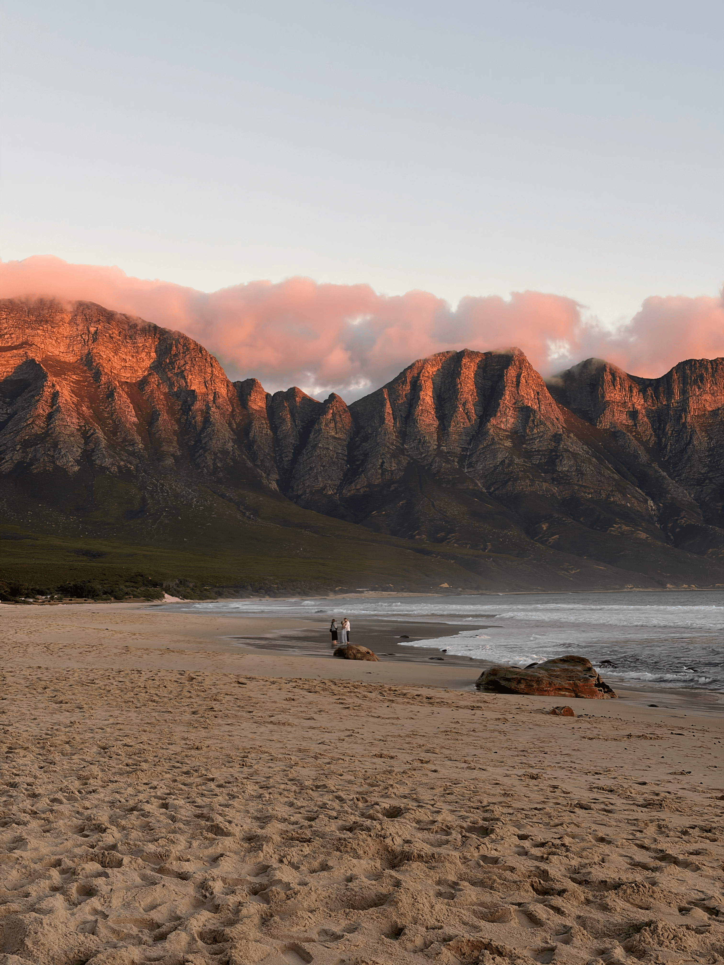 Rugged mountains glowing in sunset light overlooking a sandy beach with waves and pink clouds.