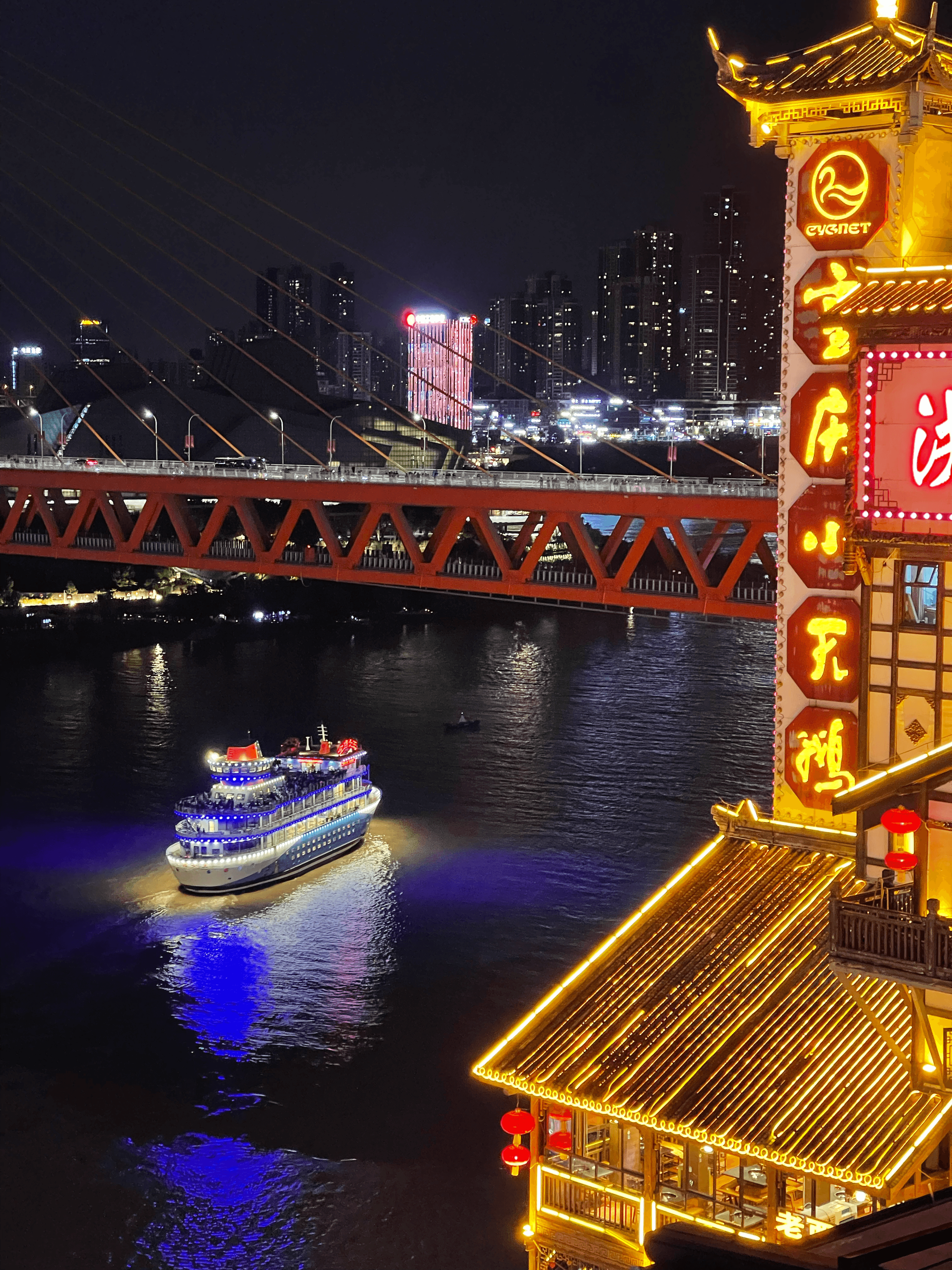 Illuminated traditional building overlooks a cruise ship and red bridge against a vibrant night cityscape.