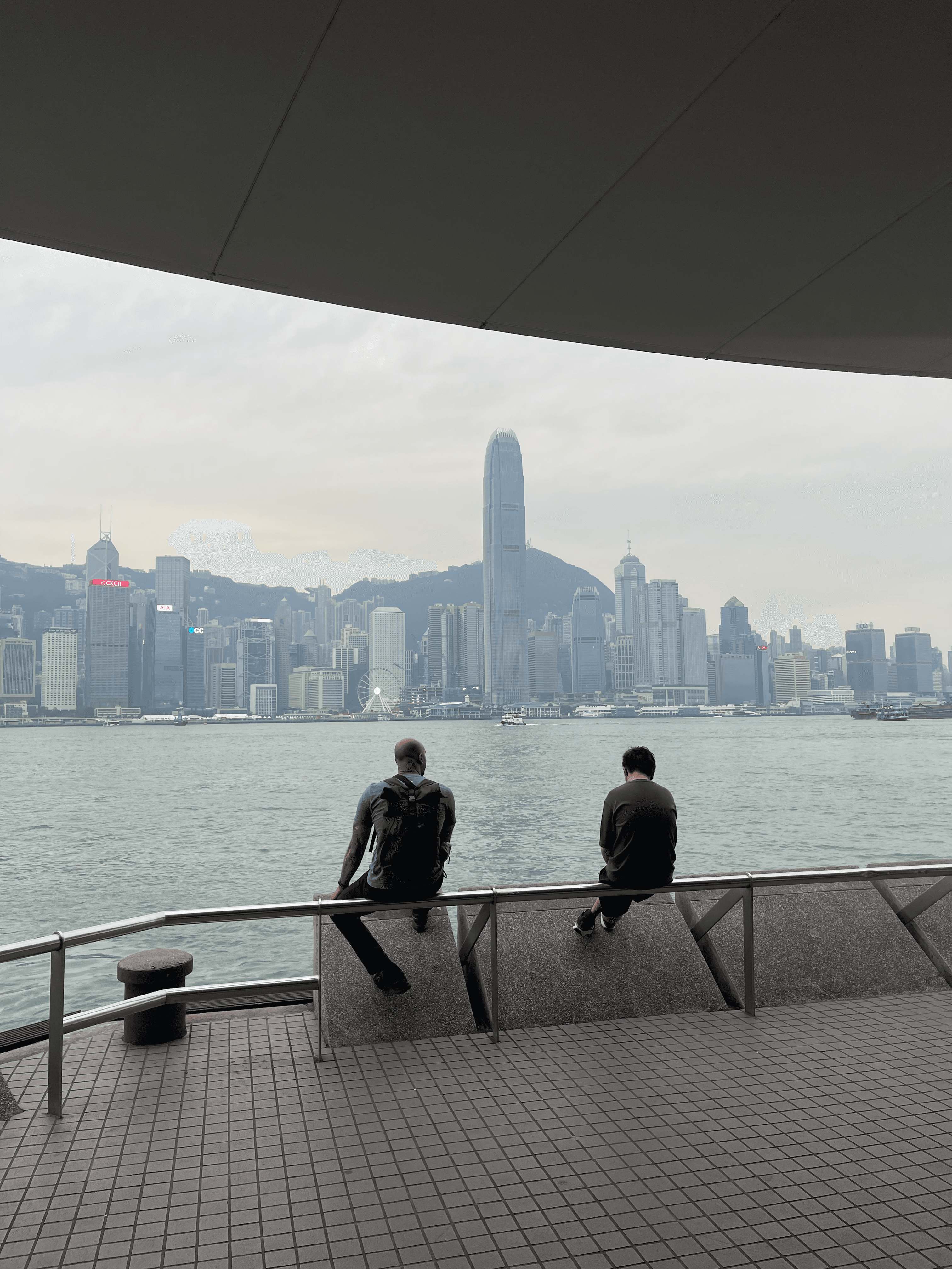 Two people sit on a railing overlooking the dense Hong Kong skyline across Victoria Harbour.